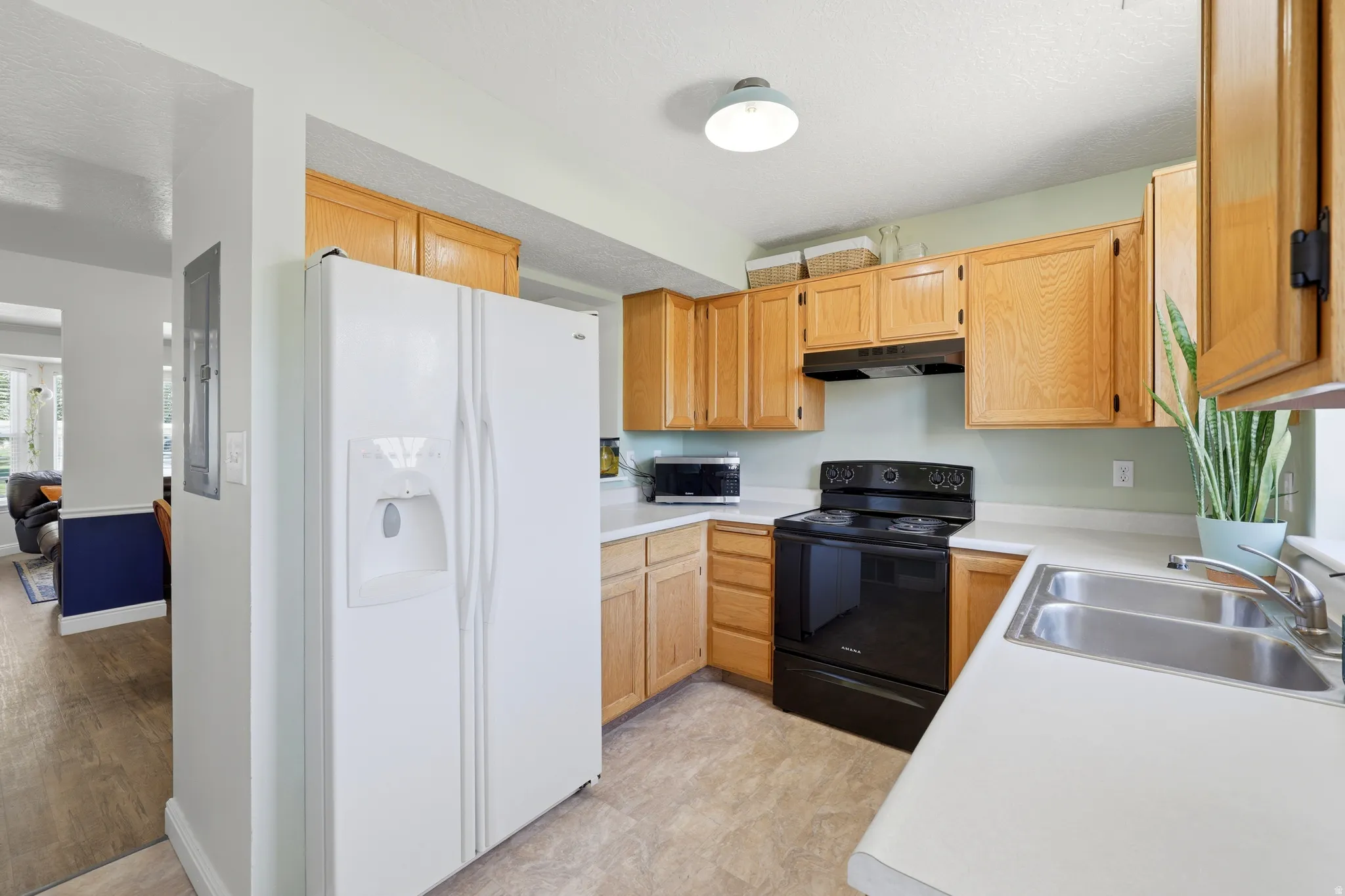 Kitchen featuring light wood cabinetry, white countertops, a double basin stainless steel sink, black electric range, and a white side-by-side refrigerator with dispenser