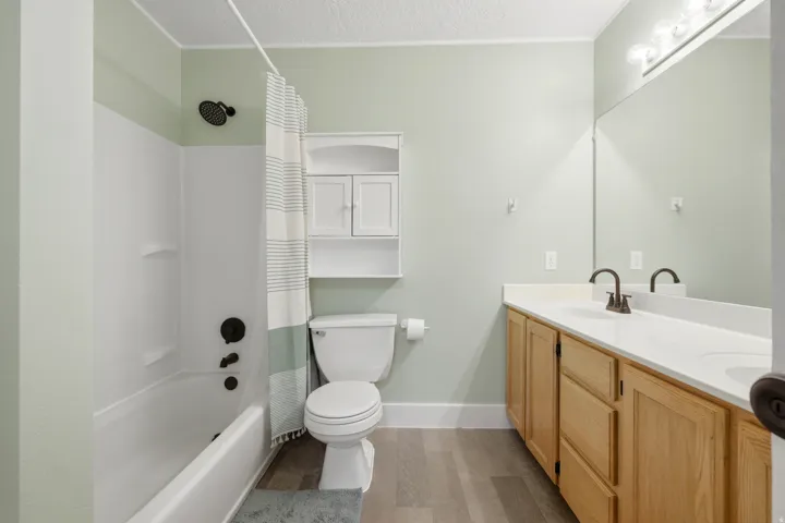 Full bathroom featuring a shower-tub combination with dark bronze fixtures, a double vanity with light wood cabinetry and a white countertop, and wood-finish flooring
