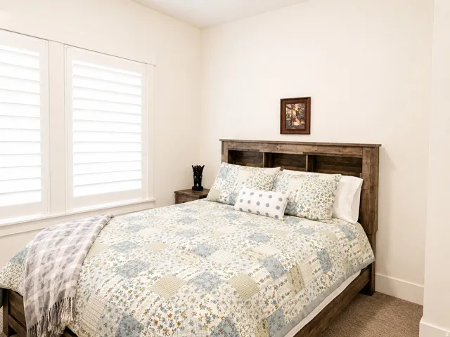 Bedroom featuring white plantation shutters, a wood-finish headboard with integrated shelving, and neutral carpeting