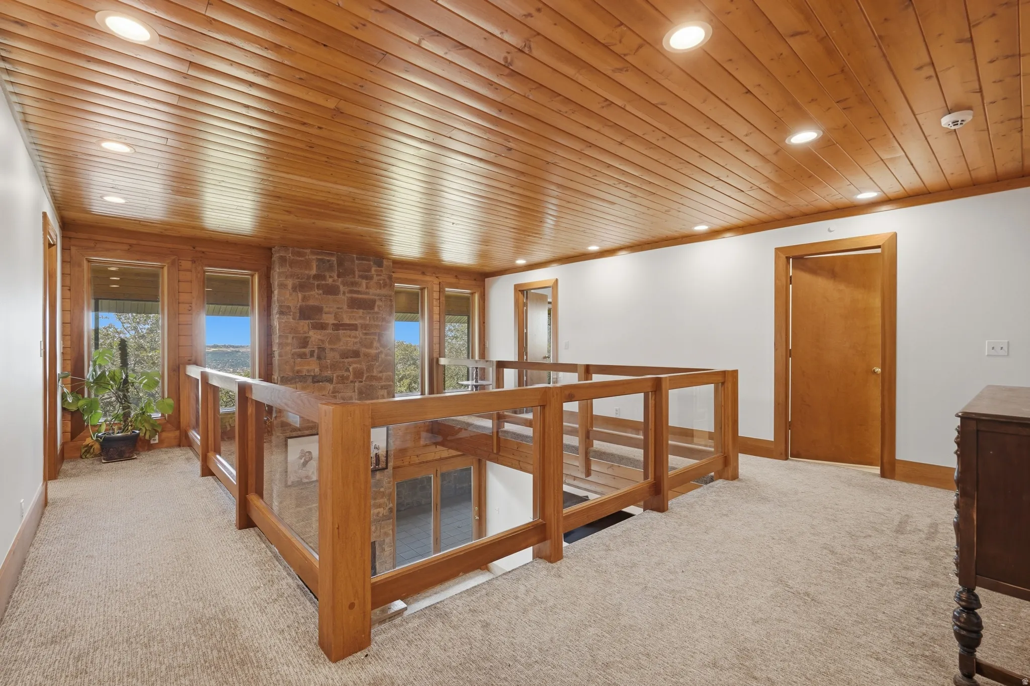 Upper-level hallway featuring a wood-panel ceiling with recessed lighting, light-toned carpet flooring, and a stone-clad wall
