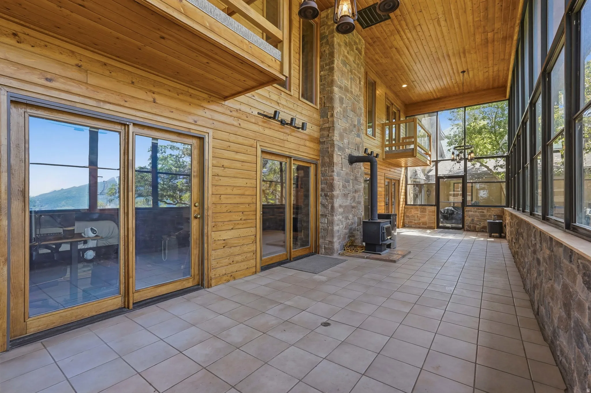 Expansive enclosed sunroom featuring a stone accent wall, wood-paneled ceiling, wood-burning stove, and extensive window-walls