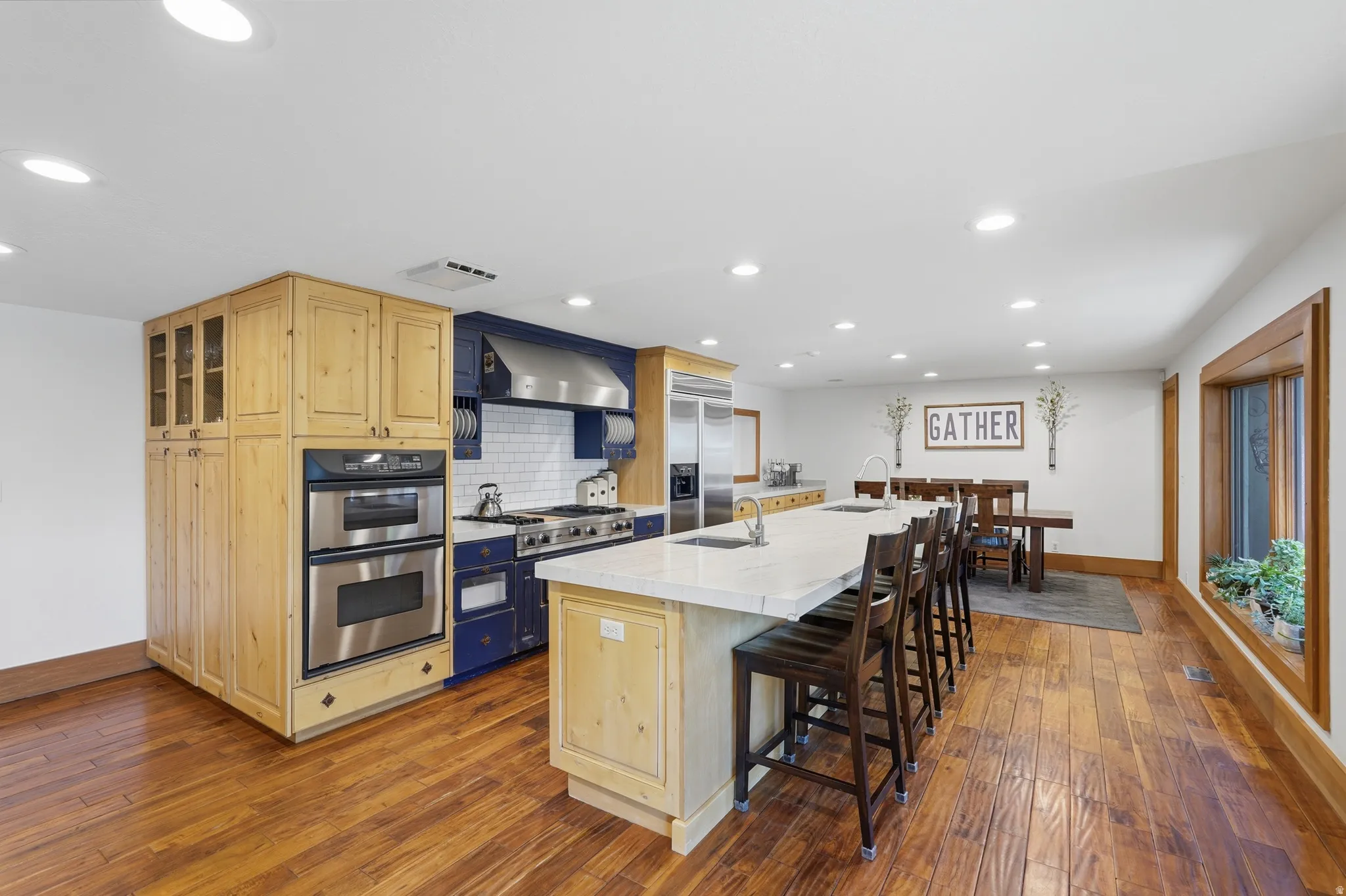 Open-concept kitchen featuring an expansive island with seating, a stainless steel gas range with a custom hood, double wall ovens, wood flooring, and recessed lighting