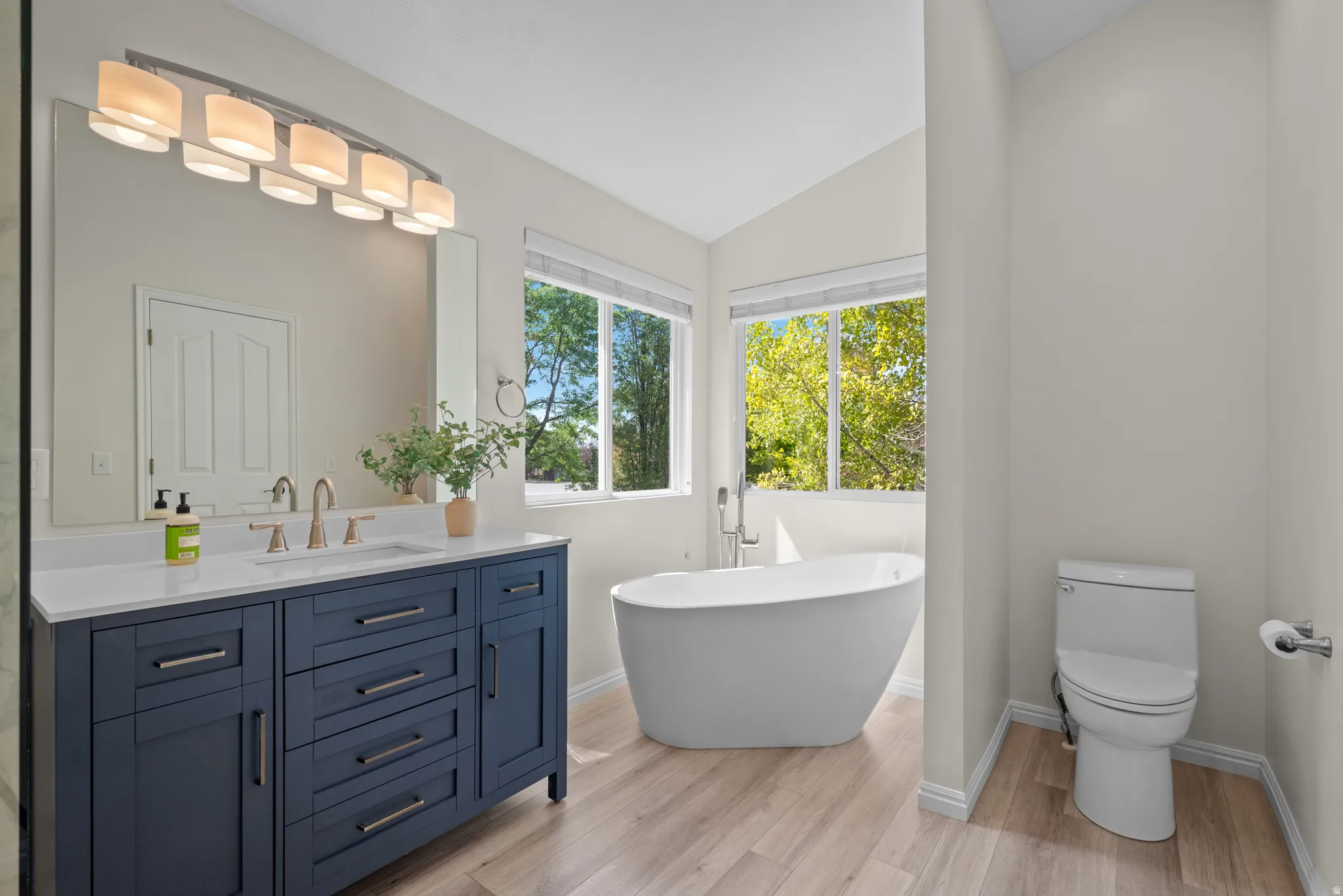 Contemporary bathroom featuring a freestanding soaking tub with floor-mounted faucet, a double vanity with a deep blue finish, a stone-look countertop, and wood-finish flooring