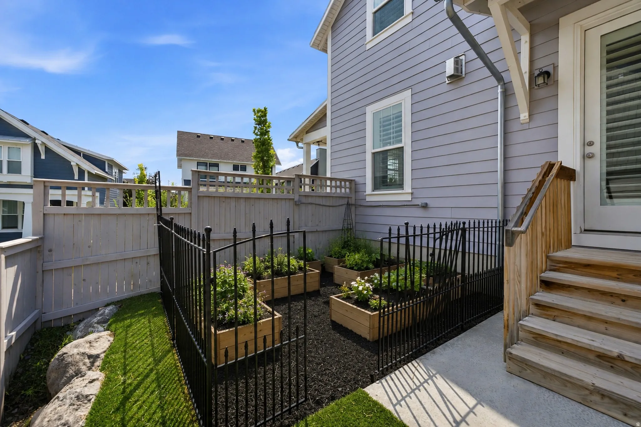 Fenced side yard with raised garden beds, artificial turf, and a concrete pathway leading to exterior wood stairs
