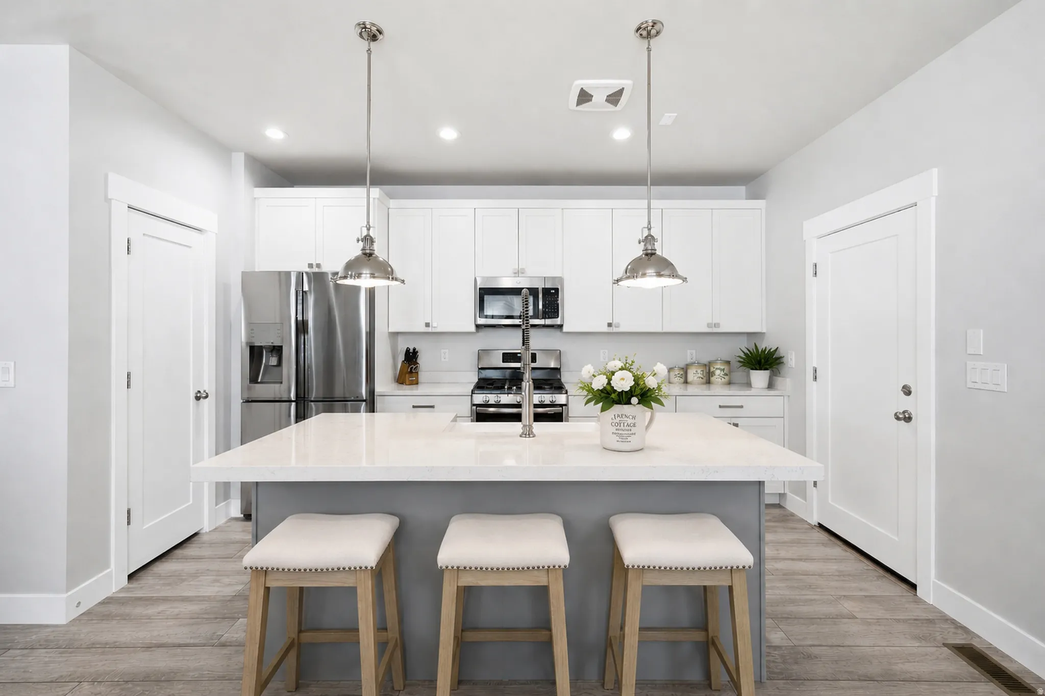 Kitchen featuring a central island with a light-toned countertop, white shaker-style cabinetry, stainless steel appliances, pendant lighting, and wood-finish flooring