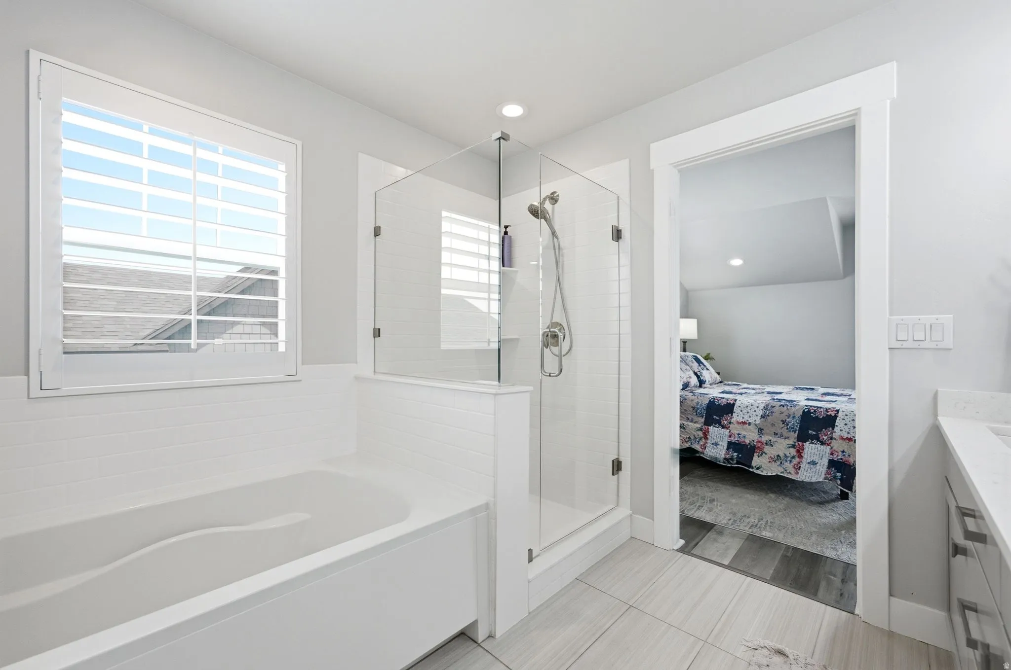 Bathroom featuring a built-in tub with white subway tile surround, a glass-enclosed shower with tile, plantation shutters, and light-toned flooring