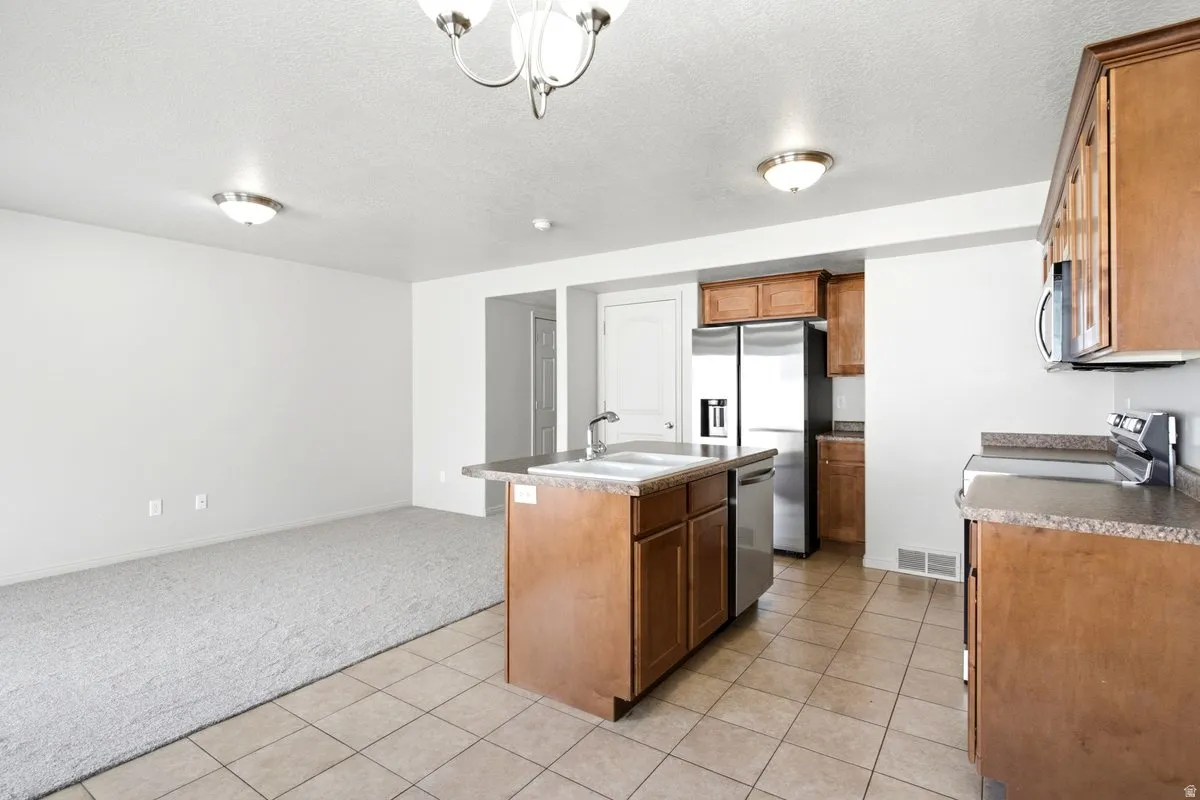 Open-concept kitchen featuring an island with a double basin sink, stainless steel appliances, wood-finish cabinetry, and tiled flooring