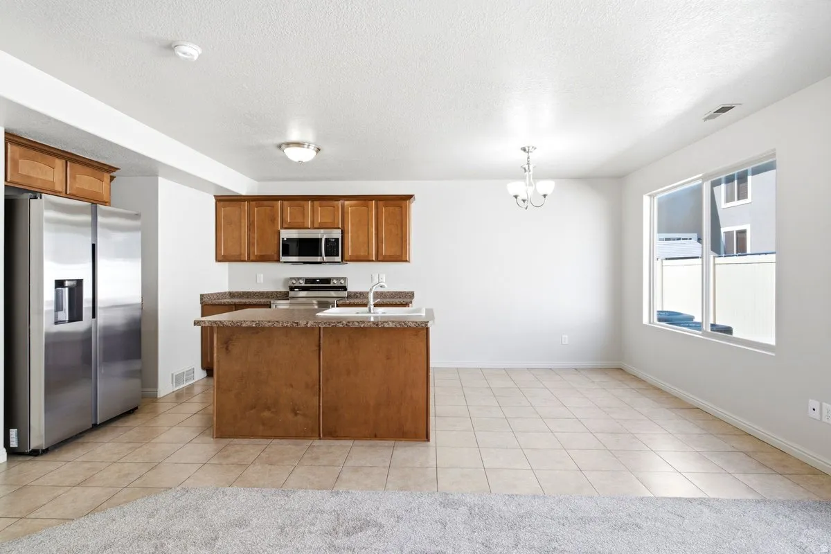 Open-concept kitchen featuring wood-finish cabinetry, stainless steel appliances, a center island with an undermount sink, and durable tile flooring