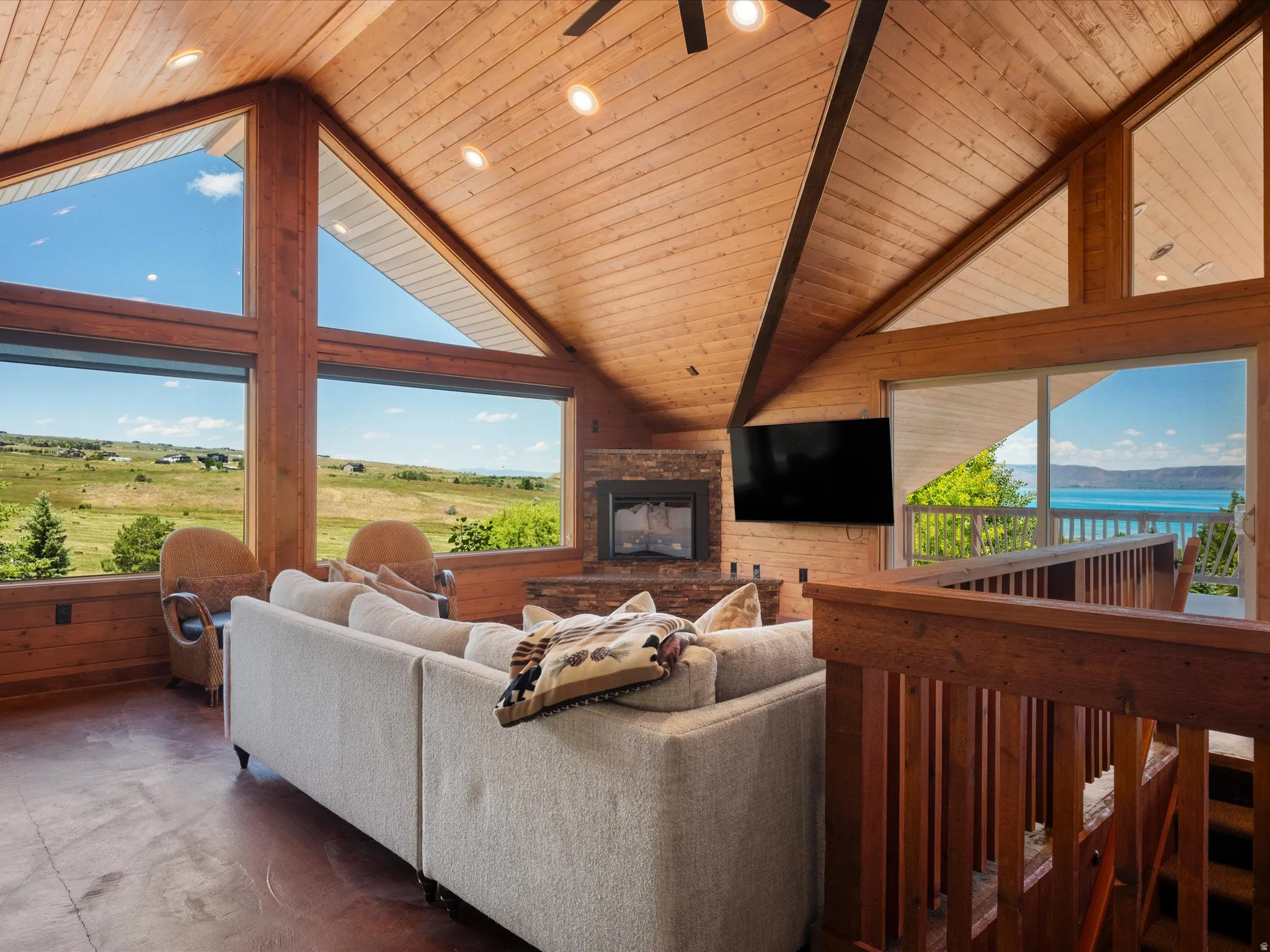 Vaulted wood-paneled ceilings with recessed lighting, floor-to-ceiling windows, a stone-faced fireplace, a sliding glass door, and wood railings