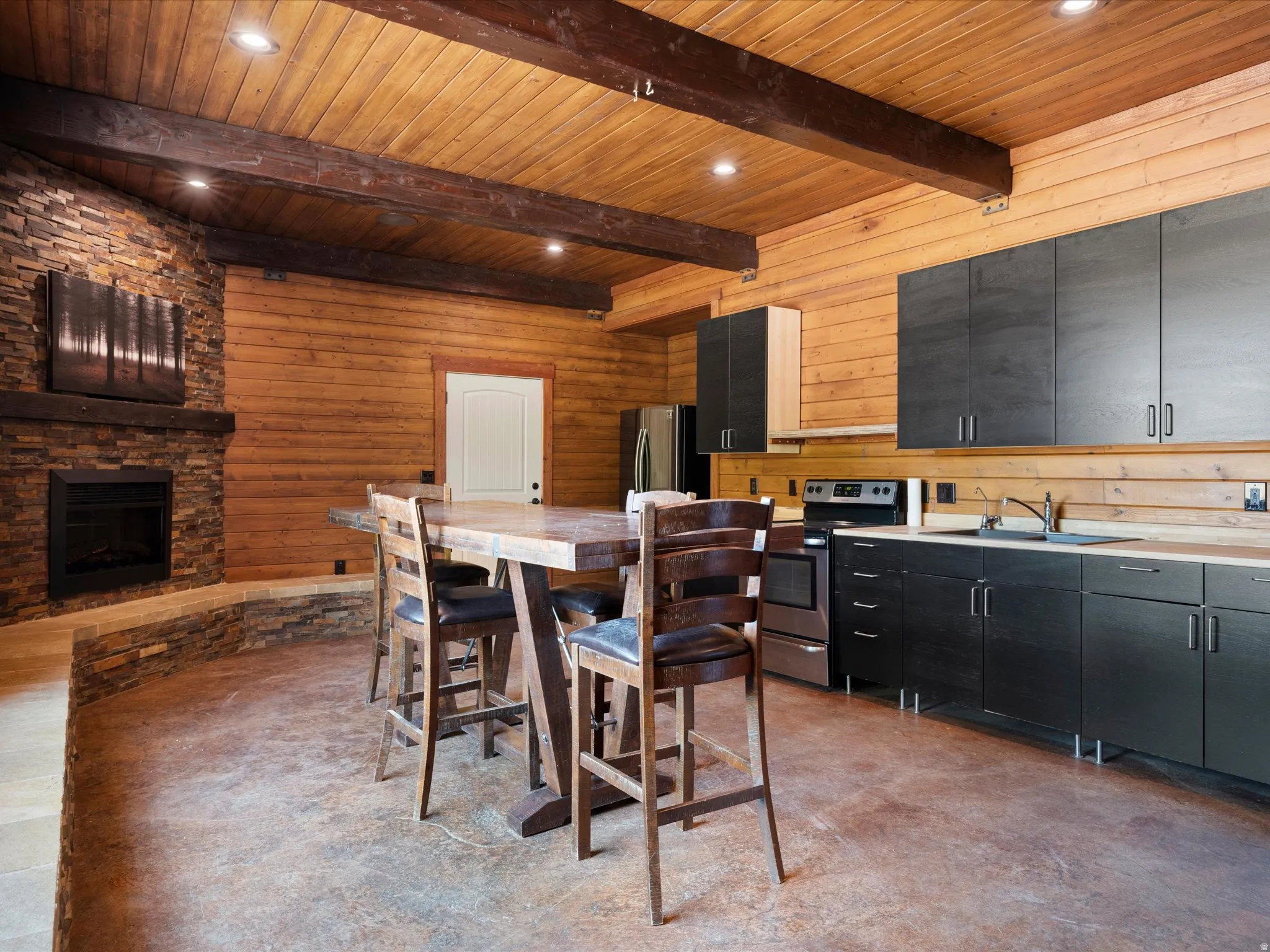 Spacious kitchen and living area featuring a stone-clad fireplace, log-style wood walls, wood-paneled ceiling with exposed beams, dark wood-finish cabinetry, and concrete flooring
