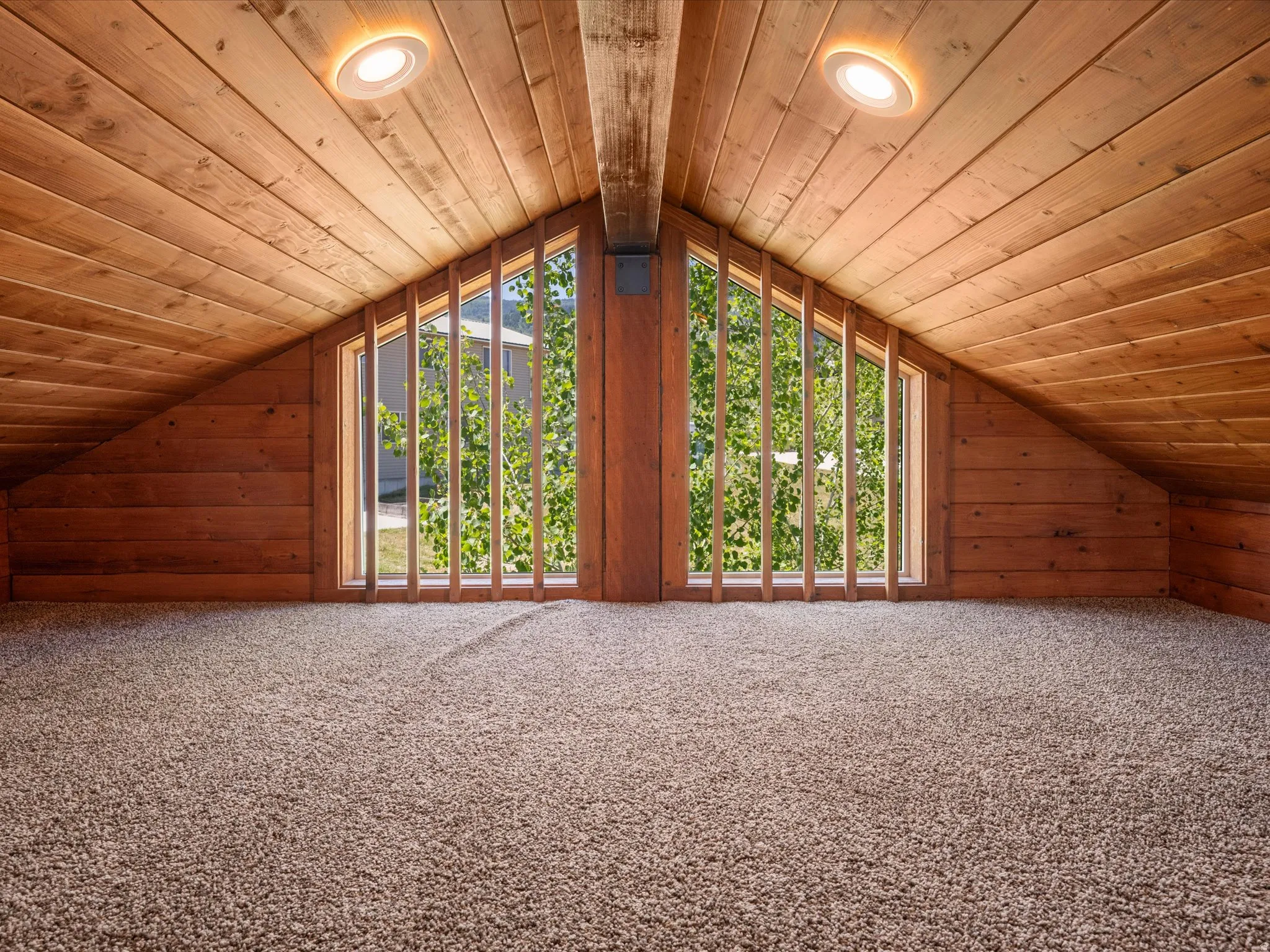 Carpeted loft area featuring a vaulted wood-paneled ceiling with exposed wood beams, two recessed ceiling lights, and twin triangular windows with vertical wood muntins