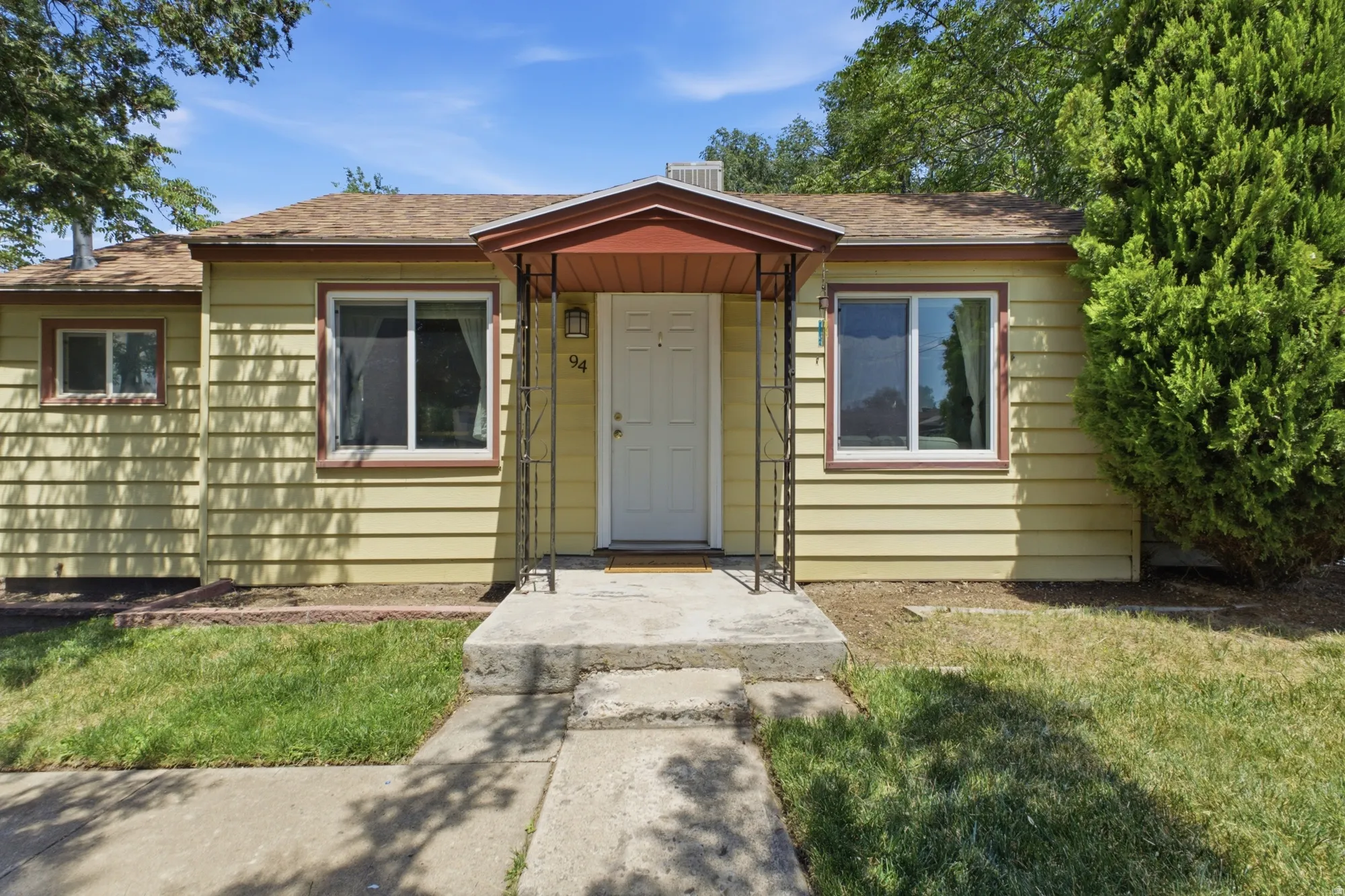 Single-story residence featuring light yellow horizontal siding, mature trees, covered entry, and welcoming curb appeal.