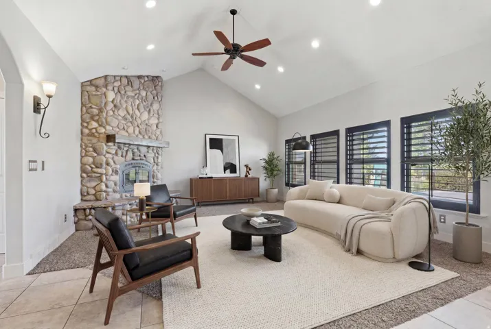 Vaulted ceiling living area featuring a prominent stone fireplace, multiple windows with dark shutters, recessed lighting, and a dark wood ceiling fan
