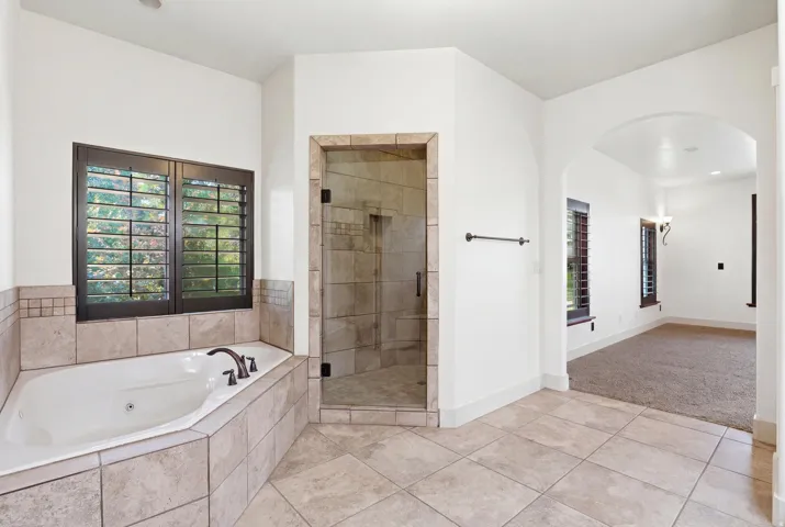 Tiled bathroom featuring a built-in soaking tub with dark hardware, a walk-in shower with clear glass enclosure, and plantation shutters