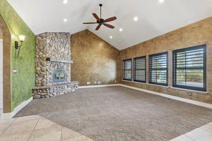 Vaulted ceiling great room featuring a stone-clad fireplace with a hearth, multiple windows with dark plantation shutters, a ceiling fan, and recessed lighting