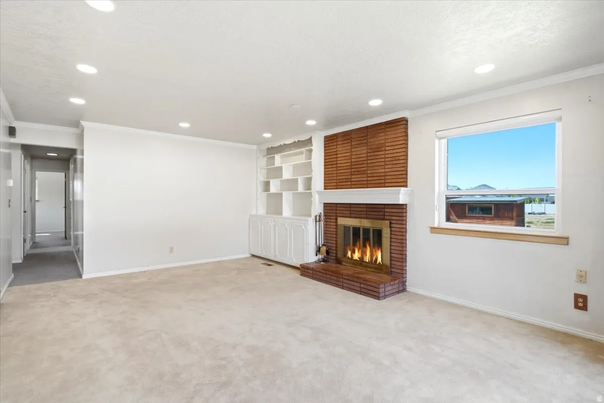 Living space featuring a brick fireplace with a white mantel, built-in shelving, recessed ceiling lights, a window, and light-toned carpet flooring