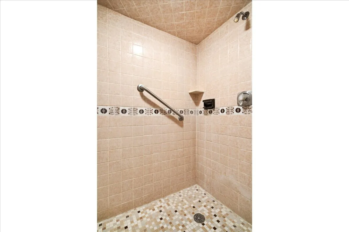 Tiled shower stall featuring mosaic floor tiling, neutral wall tiling, an accent tile band, and a chrome grab bar