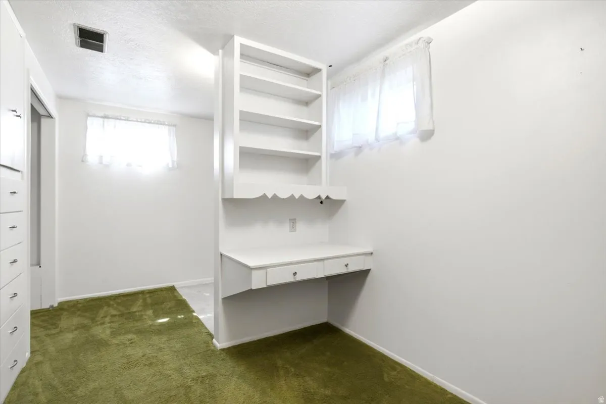 Built-in desk with shelving and integrated drawers, a window with sheer curtains, and green carpet flooring