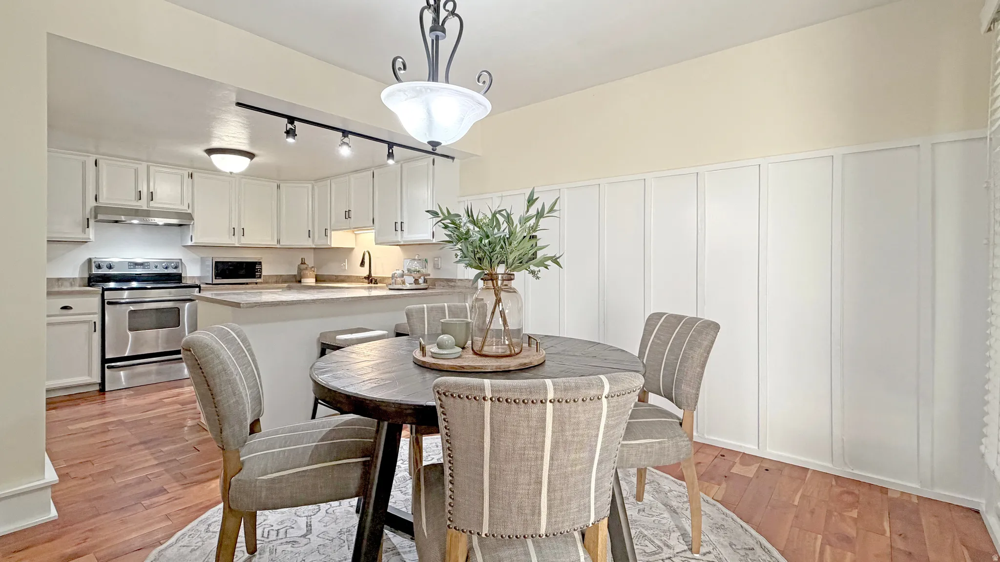 Open-concept dining area with wood-finish flooring, featuring a kitchen with white cabinetry, stainless steel appliances, and a breakfast bar