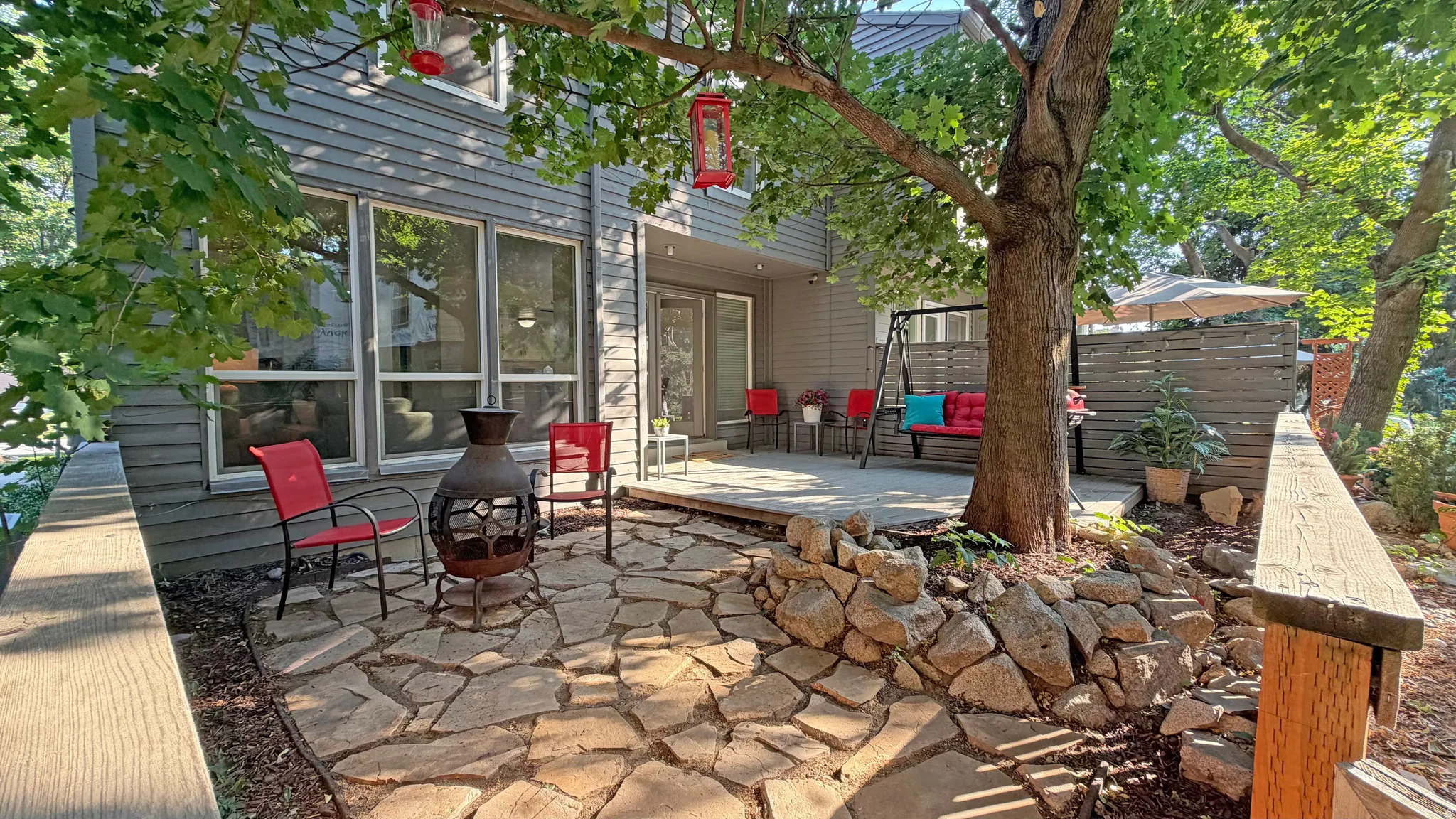 Expansive outdoor living area featuring a flagstone patio, a separate elevated deck, a stone-edged planting bed, a mature shade tree, and a slatted privacy fence