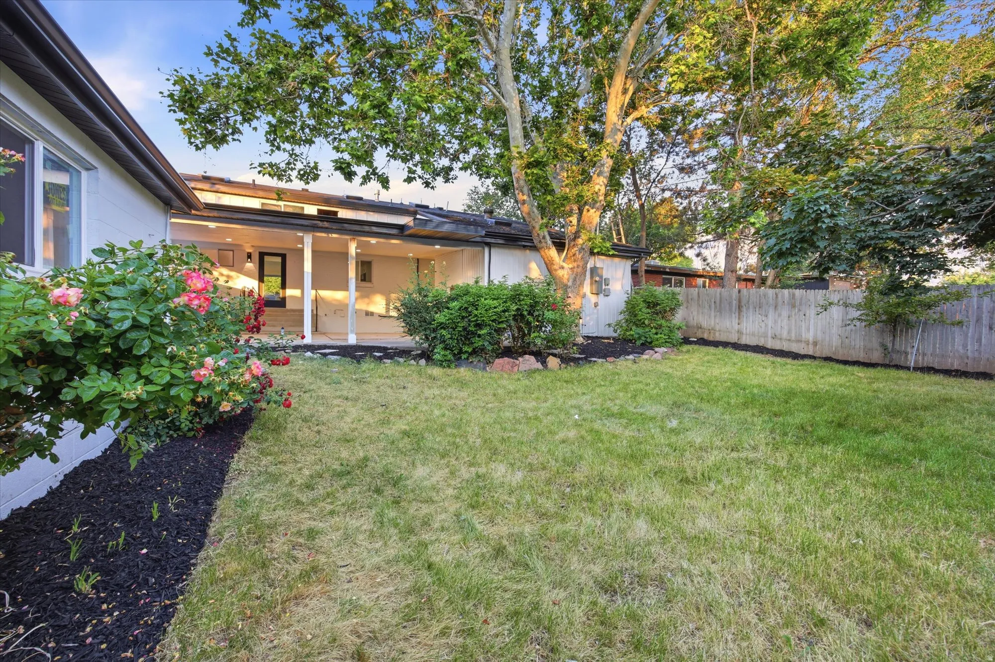 Expansive rear yard featuring a covered patio, privacy fencing, mature trees, and landscaping with flowering shrubs