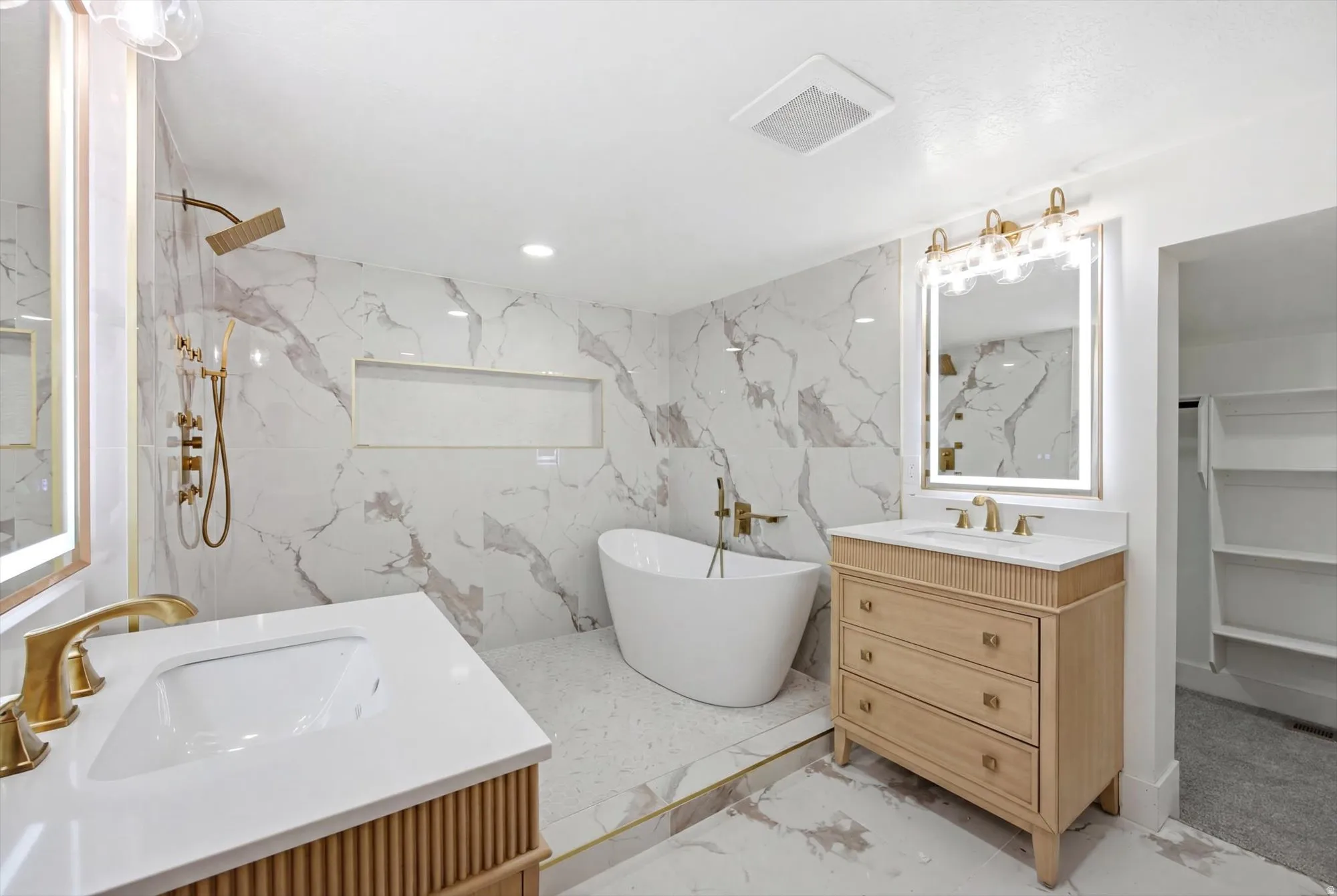 Bathroom featuring marble-patterned tile, a freestanding soaking tub, and dual vanity sinks with gold-tone fixtures