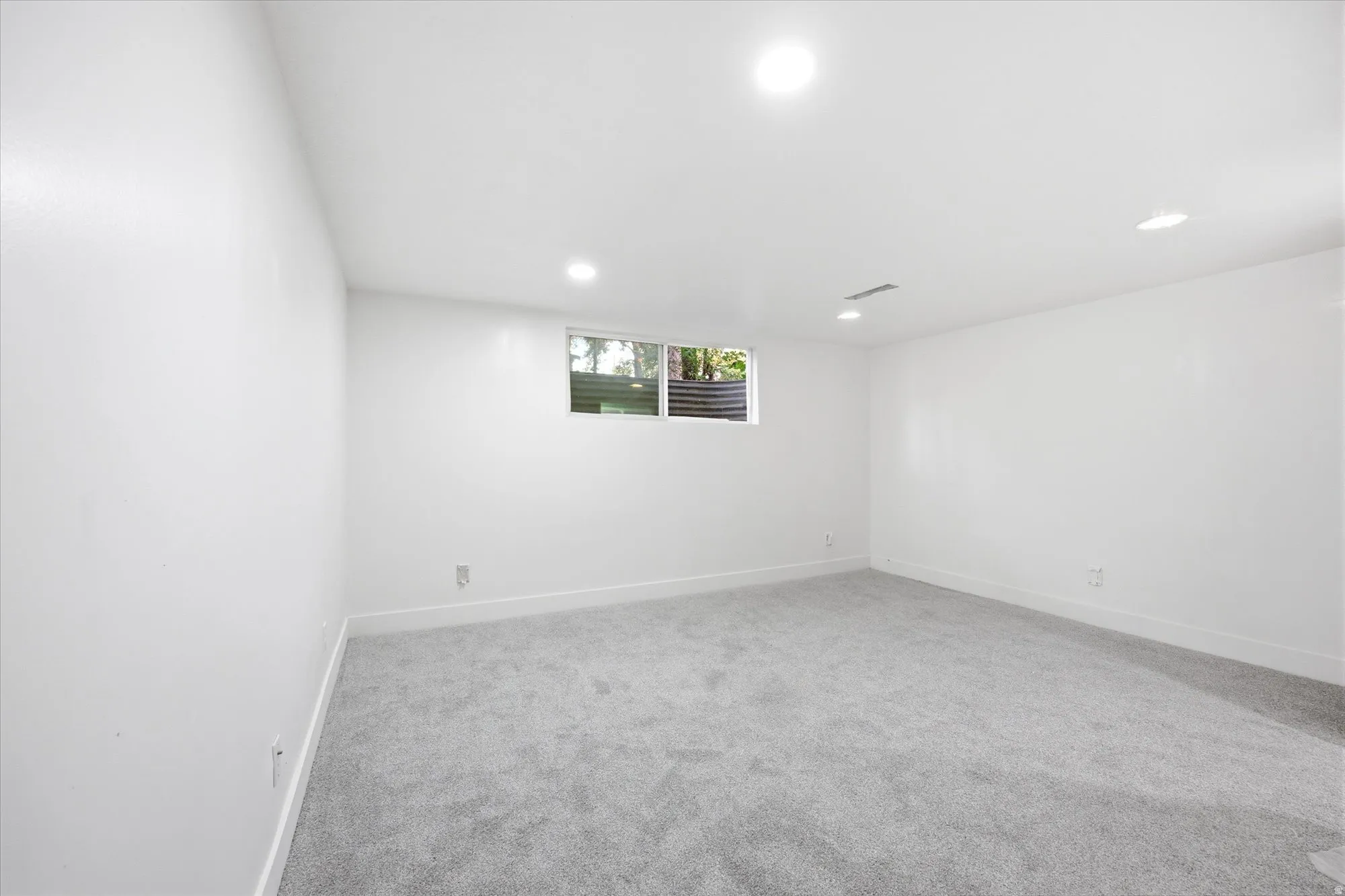 Spacious interior room featuring light gray carpet flooring, white walls, recessed ceiling lighting, and a single window