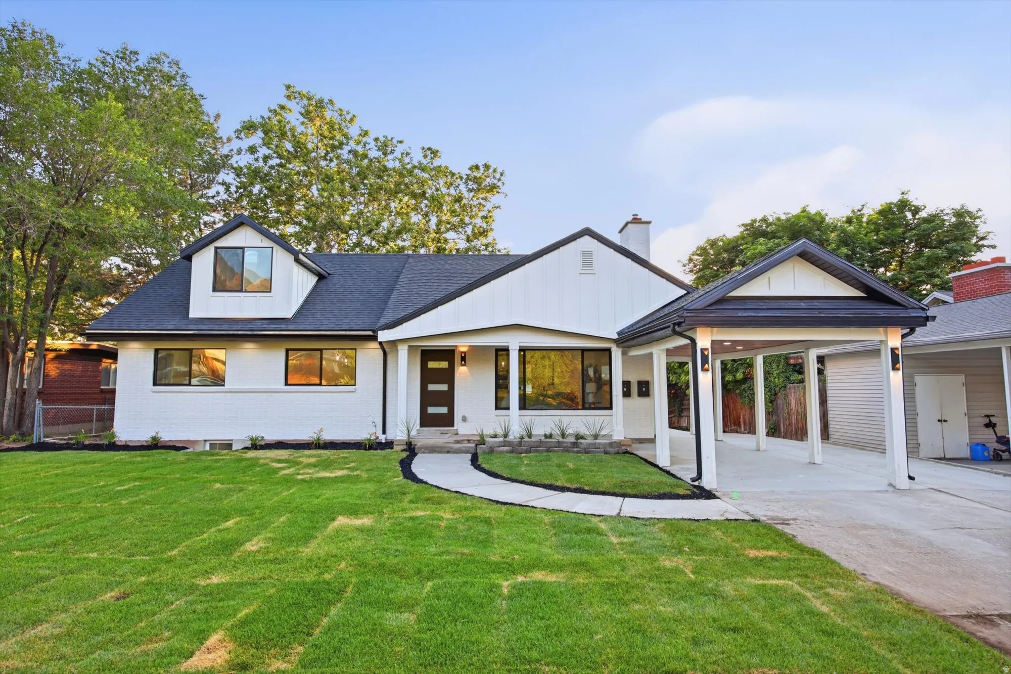 Modern-style residence featuring a carport, dark shingle roof, white board-and-batten siding, black-framed windows, and a concrete pathway leading to the front entry