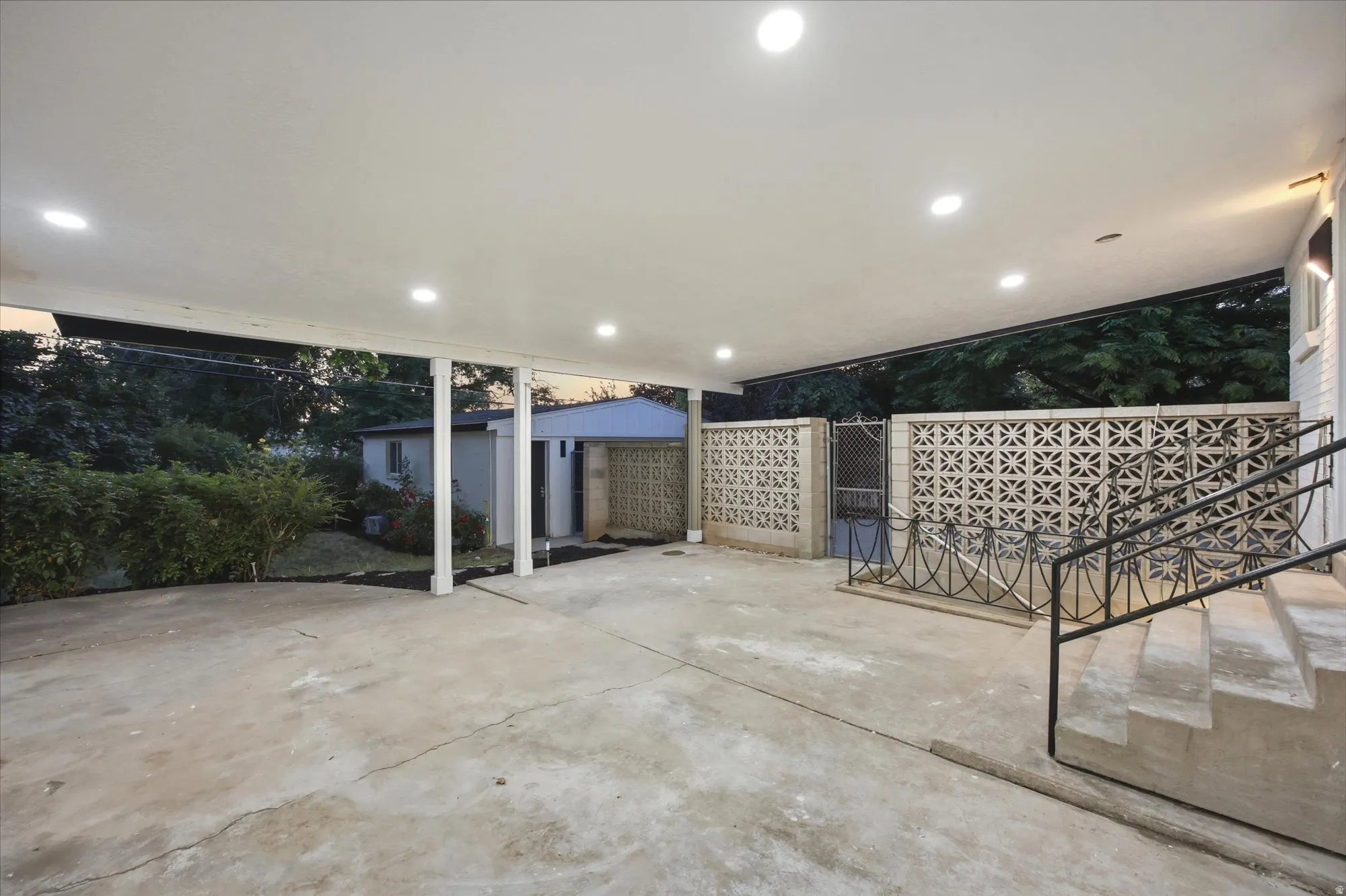 Covered outdoor patio area featuring recessed lighting, a concrete surface, and wrought iron railings leading to an elevated entrance