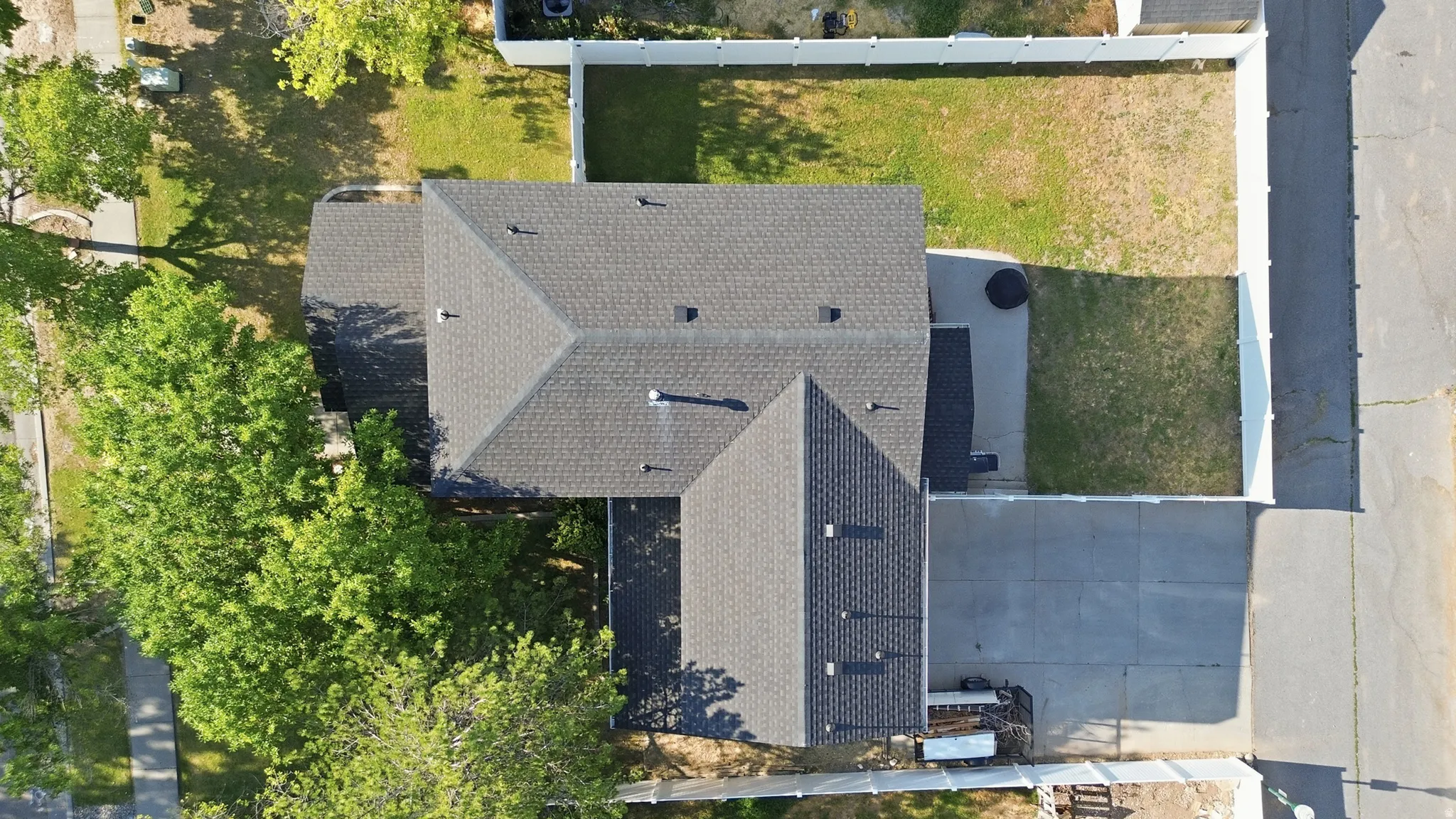 Aerial perspective showcasing a property with a dark shingle roof, a concrete driveway, and a fenced backyard featuring a patio area