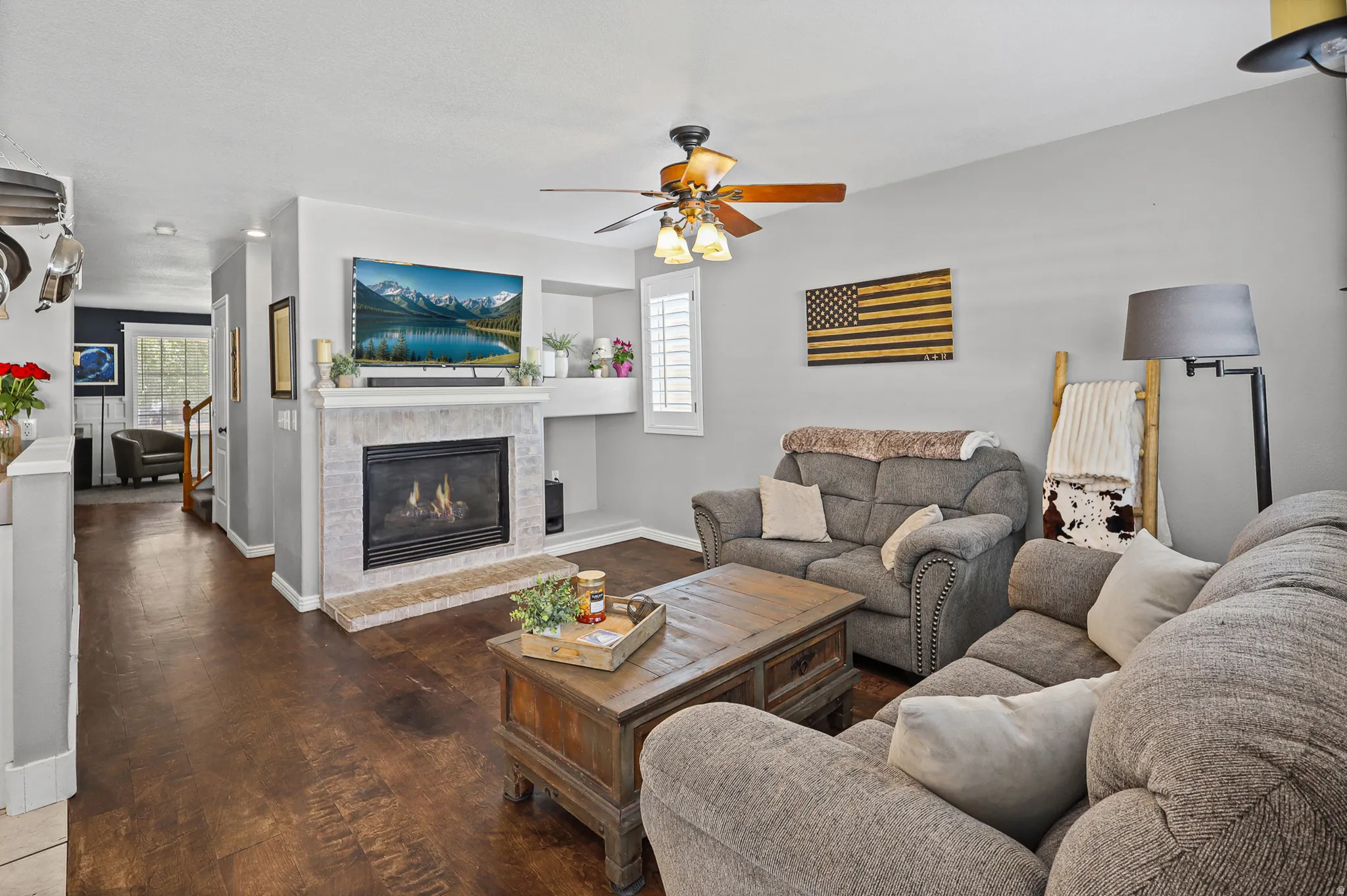 Living area featuring wood-finish flooring, a tiled fireplace with a mantel, a ceiling fan with integrated lighting, and white plantation shutters