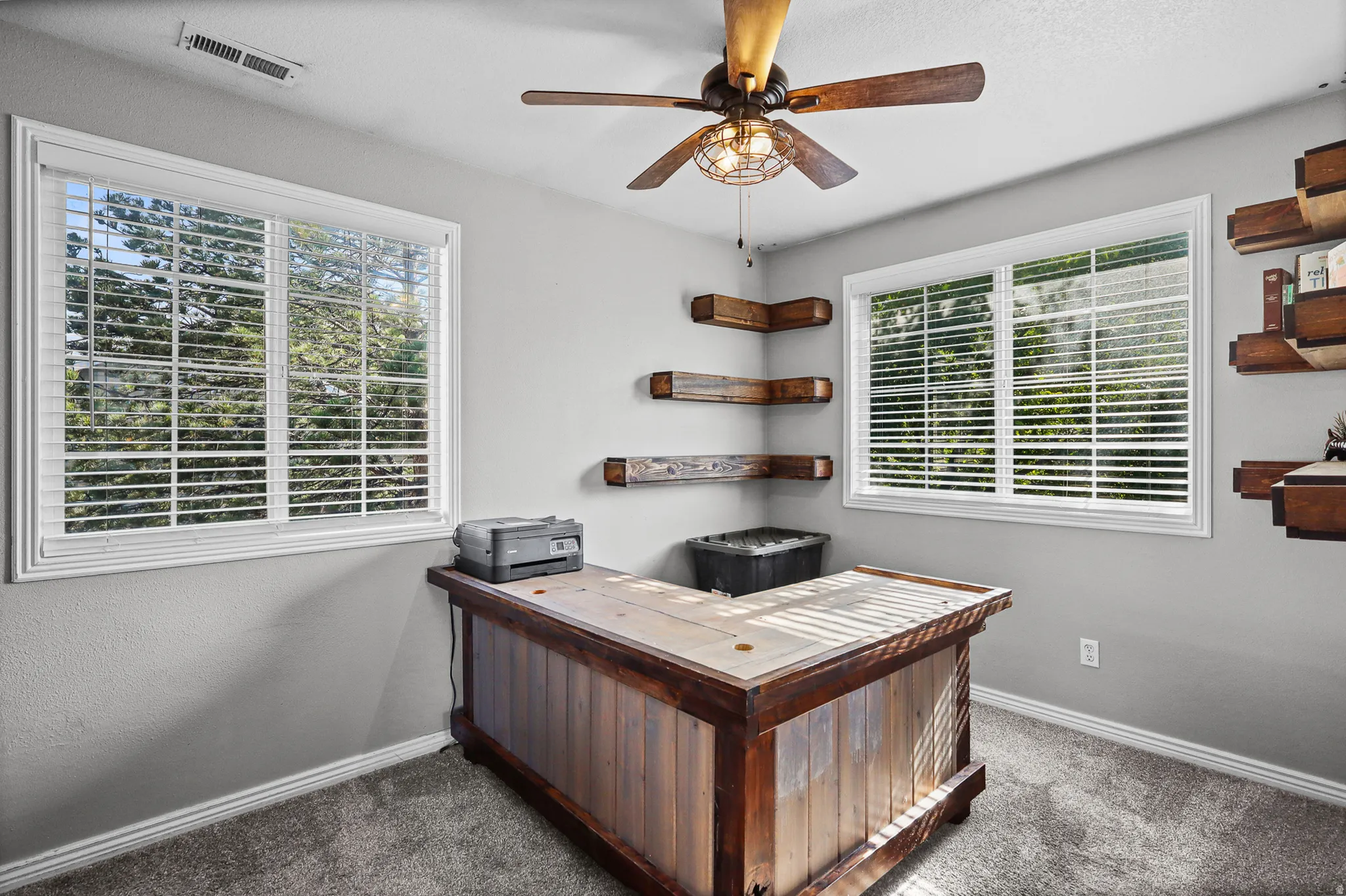 Bright room featuring two windows with white blinds, a ceiling fan with a decorative light fixture, and wood-finish floating corner shelves