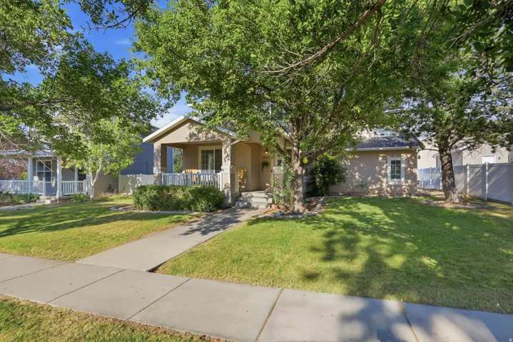 Stucco exterior residence featuring a covered front porch with white railing, established front yard with mature trees, paved sidewalk, and a partial privacy fence