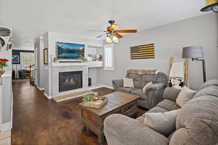 Living area featuring wood-finish flooring, a tiled fireplace with a mantel, a ceiling fan with integrated lighting, and white plantation shutters
