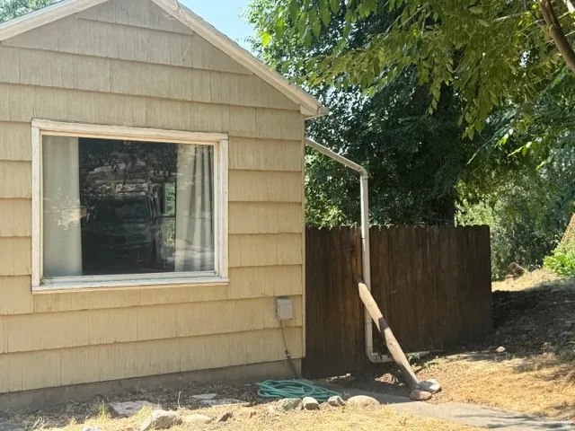 Beige shingle siding exterior featuring a rectangular window with white trim, a brown wood fence, and mature trees in the background