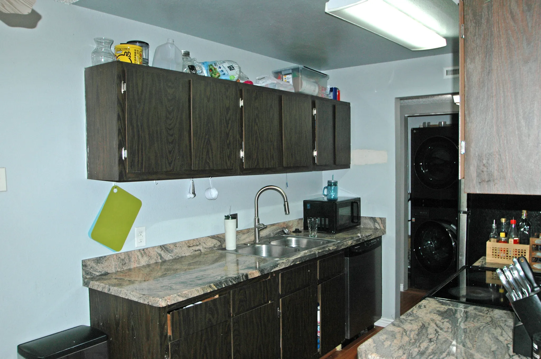 Kitchen featuring dark wood-finish cabinetry, a double basin stainless steel sink with a gooseneck faucet, a built-in dishwasher, and stone-look countertops