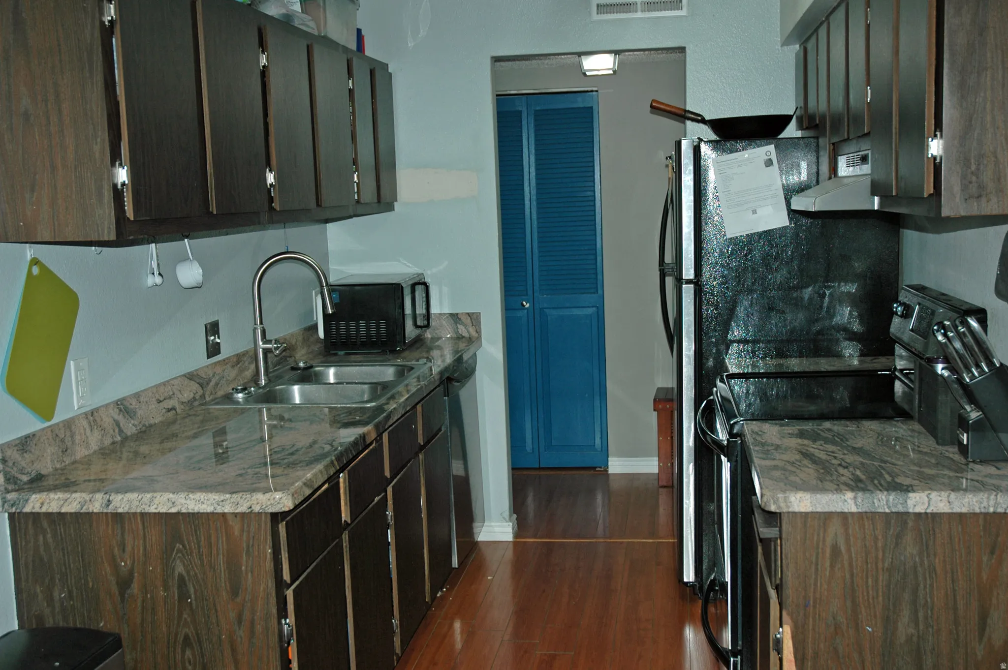 Galley kitchen featuring wood-finish cabinetry, granite-style countertops, stainless steel double basin sink, and wood-finish flooring
