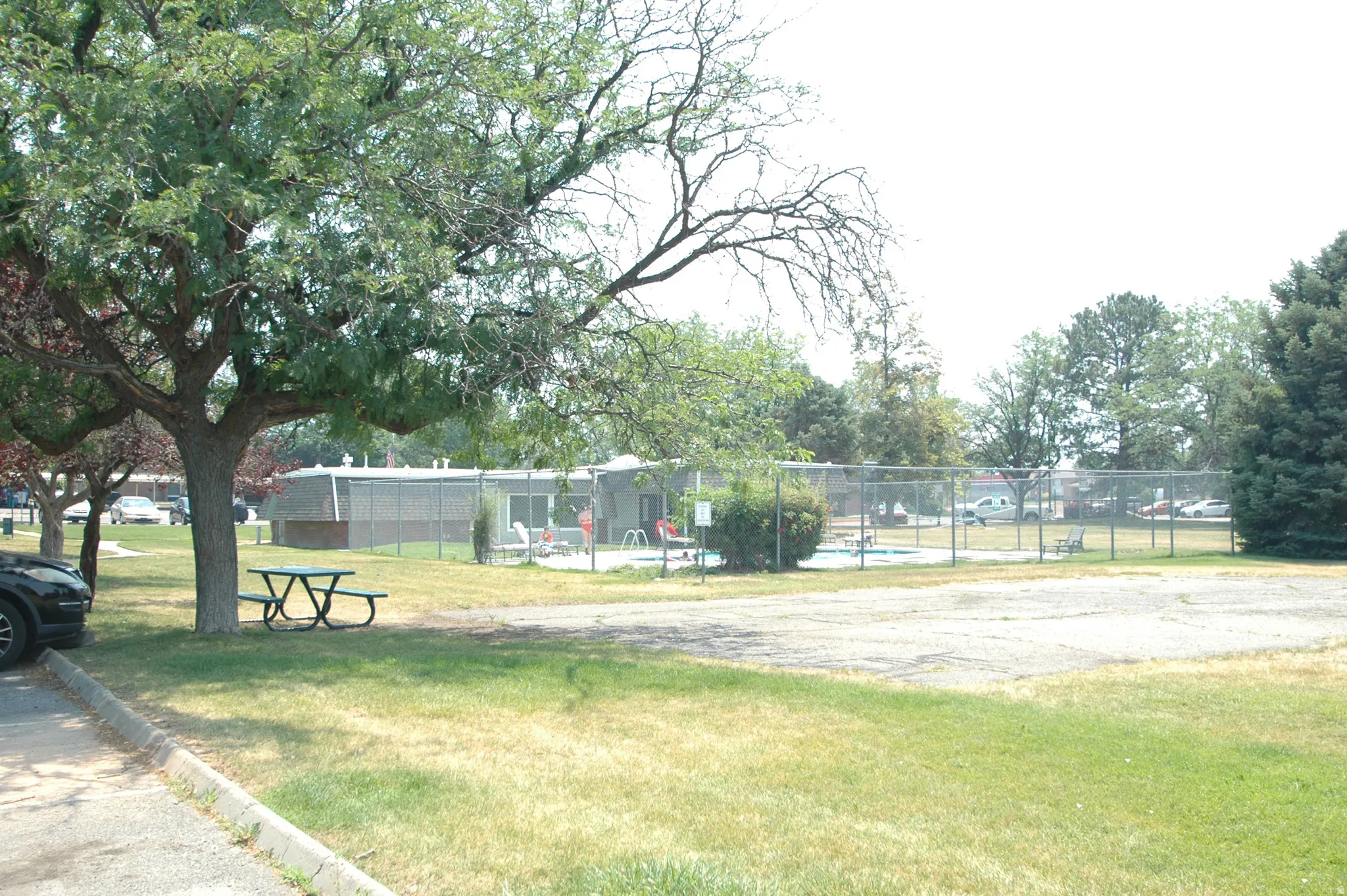 Recreational area featuring a fenced swimming pool, surrounding green space, mature trees, and a picnic table