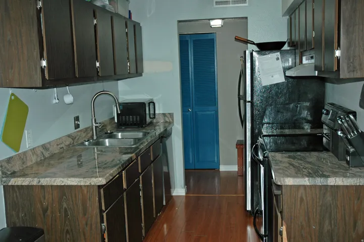 Galley kitchen featuring wood-finish cabinetry, granite-style countertops, stainless steel double basin sink, and wood-finish flooring