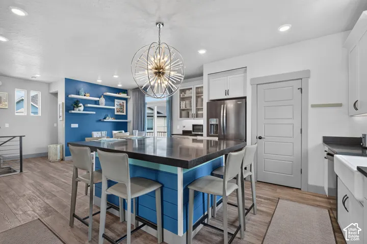 Large kitchen island with seating, a contemporary chandelier, stainless steel refrigerator, white shaker cabinetry, and wood-finish flooring