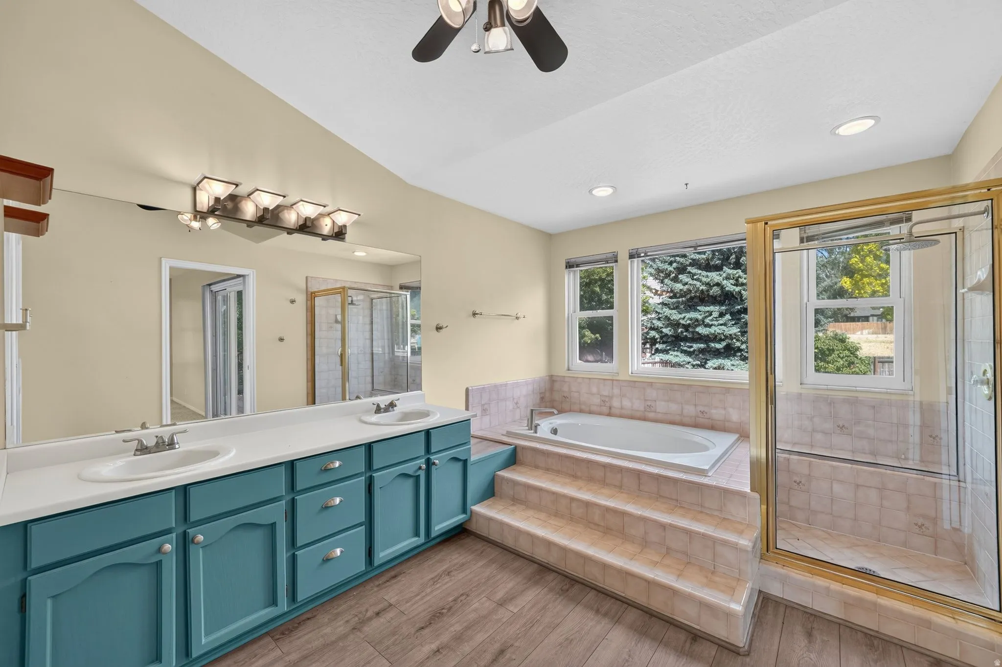 Spacious bathroom featuring an integrated soaking tub with tiled surround, a separate glass-enclosed shower, dual vanity with teal cabinetry and white countertops, and wood-finish flooring