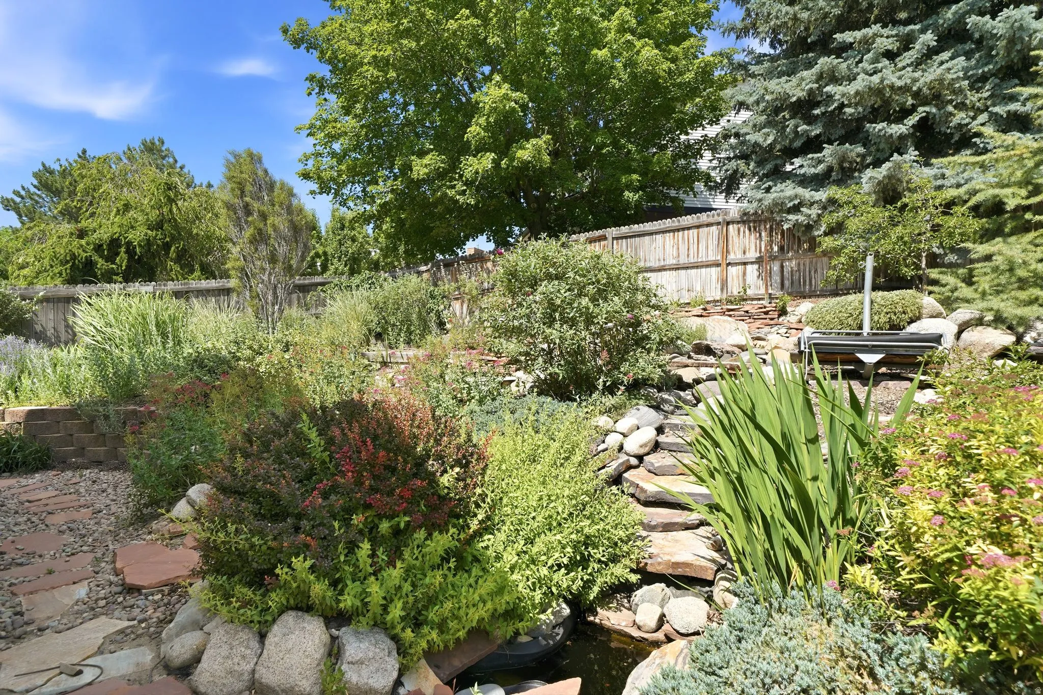 Tiered landscaping featuring a stone water feature, mature evergreens, diverse shrubbery, and a flagstone pathway