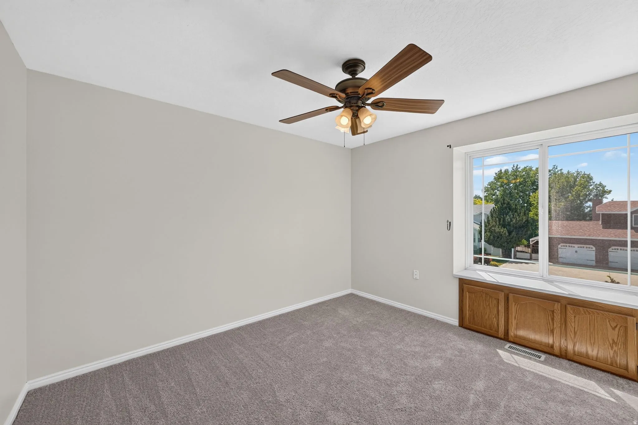 Carpeted interior space featuring a large window with white trim, built-in wooden cabinetry, a ceiling fan with integrated lighting, and neutral-toned walls