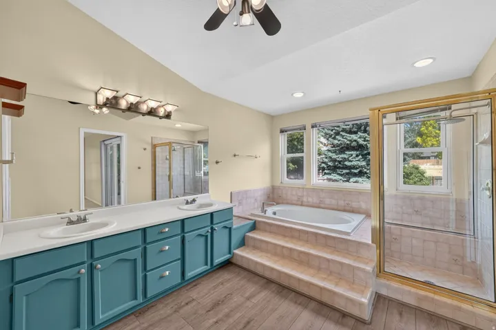 Spacious bathroom featuring an integrated soaking tub with tiled surround, a separate glass-enclosed shower, dual vanity with teal cabinetry and white countertops, and wood-finish flooring