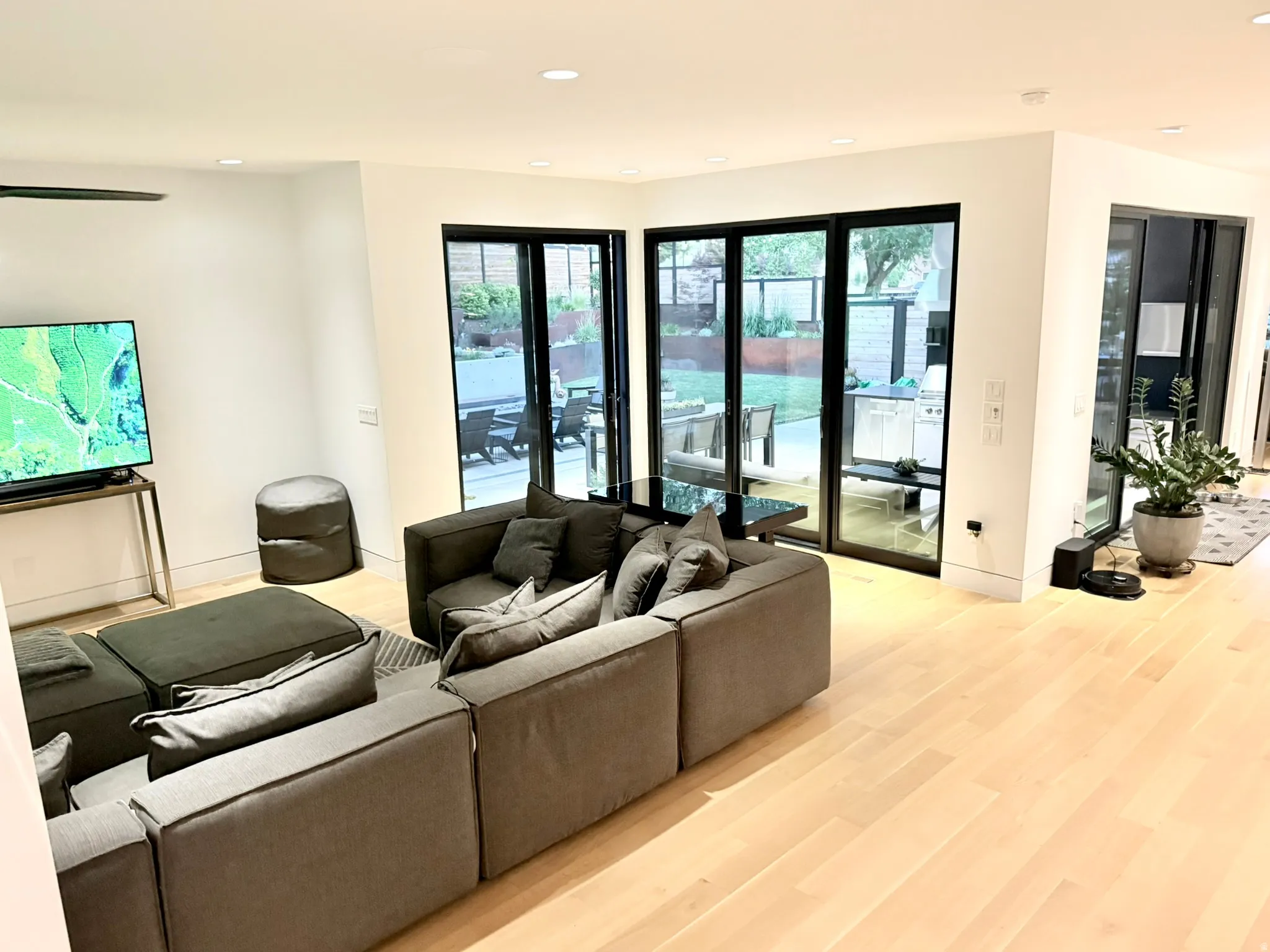 Open-concept living space with light wood-finish flooring, recessed lighting, and expansive glass doors opening to an outdoor patio with built-in barbecue area