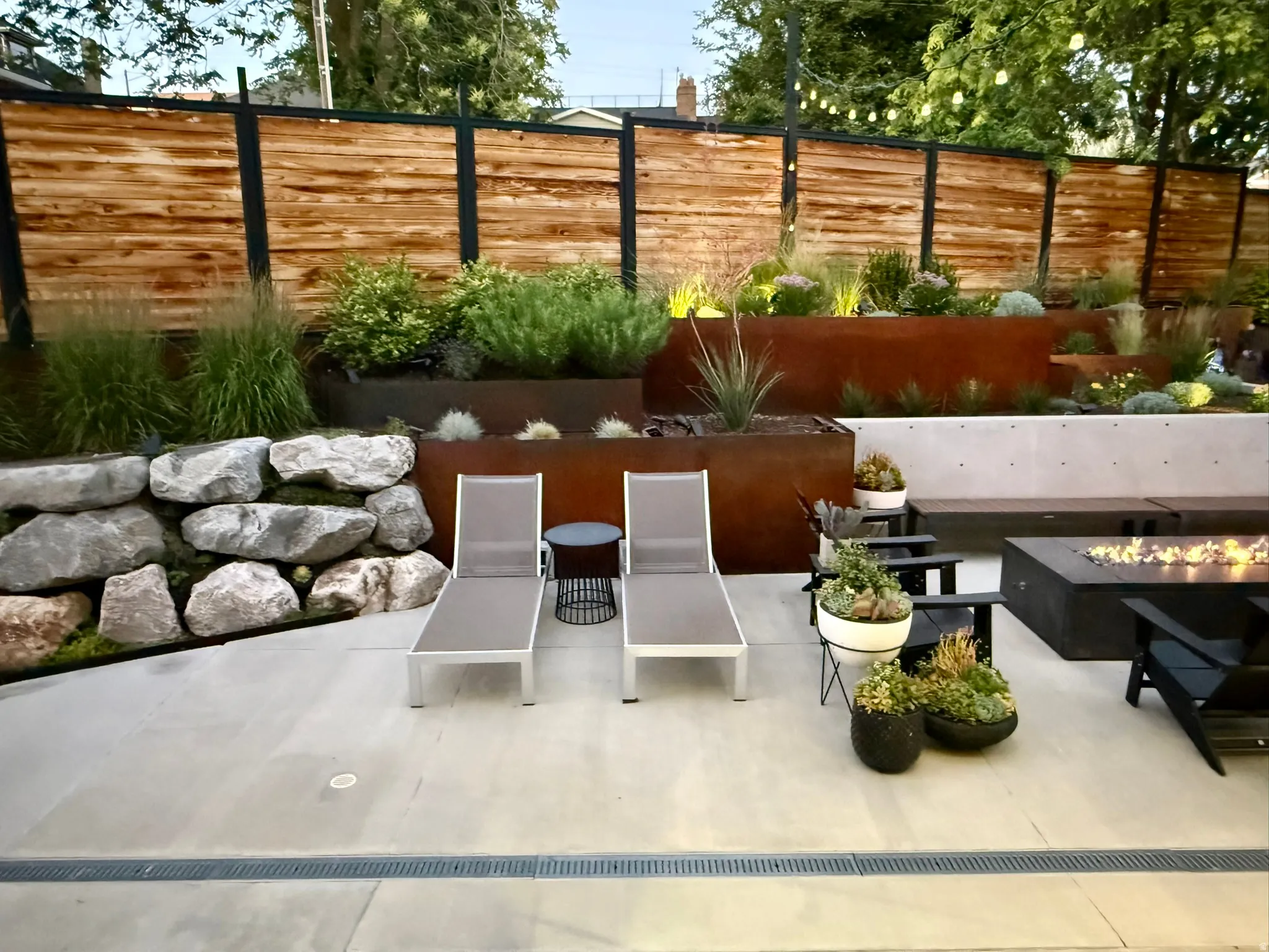 Concrete patio featuring a linear drain system, a stone retaining wall, and a slatted wood fence with black metal posts