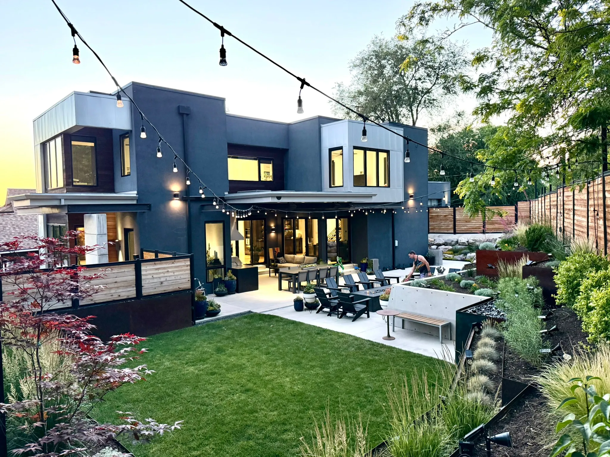 Contemporary exterior with a multi-level design featuring dark gray and light gray siding, expansive patio spaces, a manicured lawn, and terraced landscaping with mature plantings