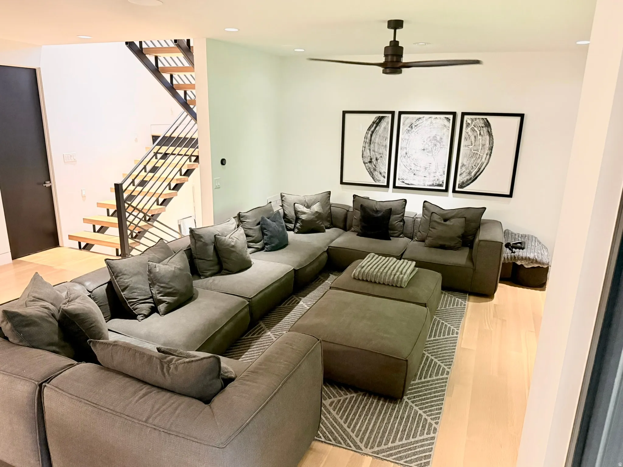 Modern living area featuring light wood-finish flooring, a contemporary ceiling fan, recessed lighting, and a floating staircase with dark metal railings
