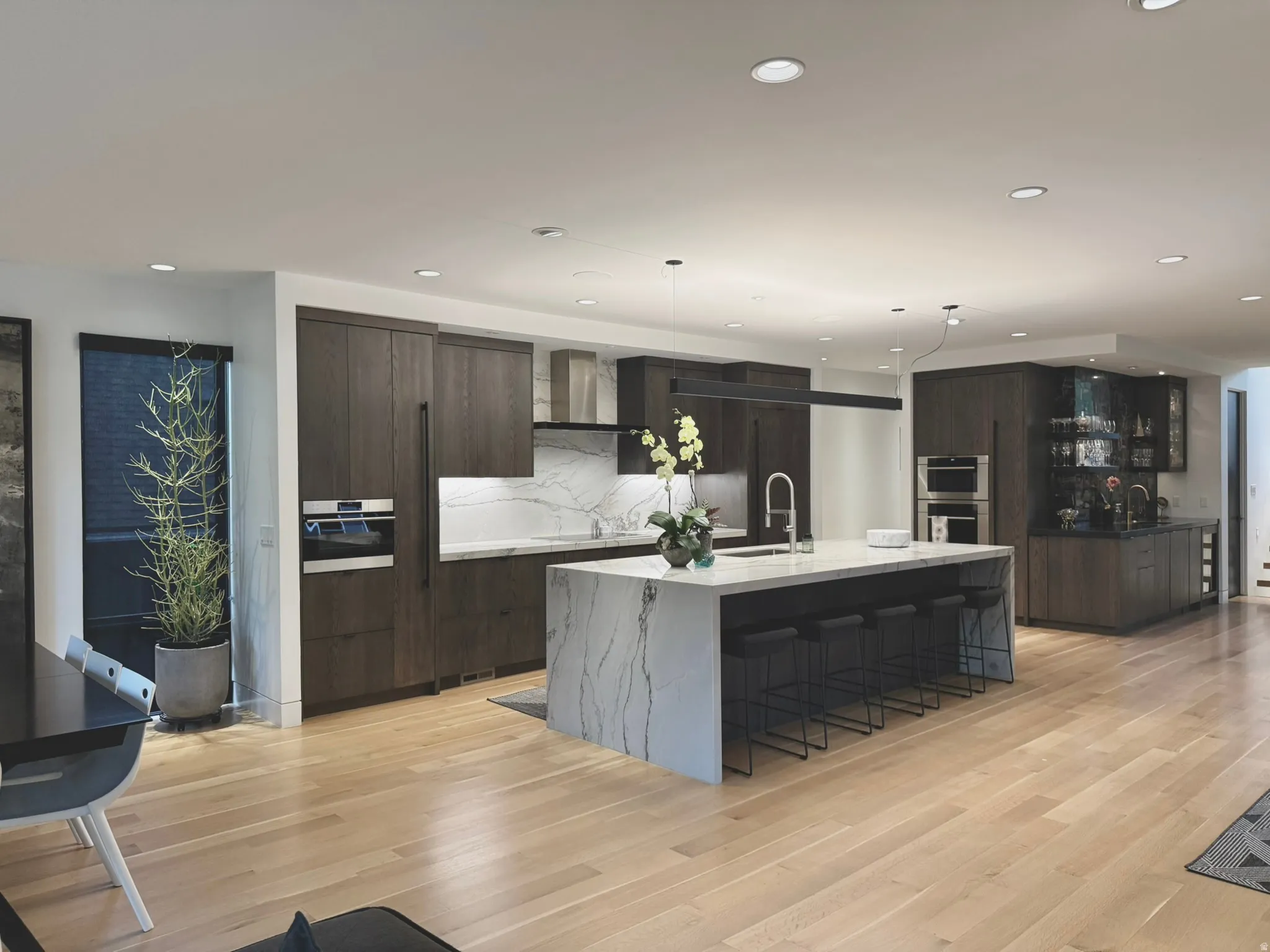 Gourmet kitchen featuring a waterfall island, custom dark wood cabinetry, integrated stainless steel appliances, marble-look countertops and backsplash, and light wood-finish flooring