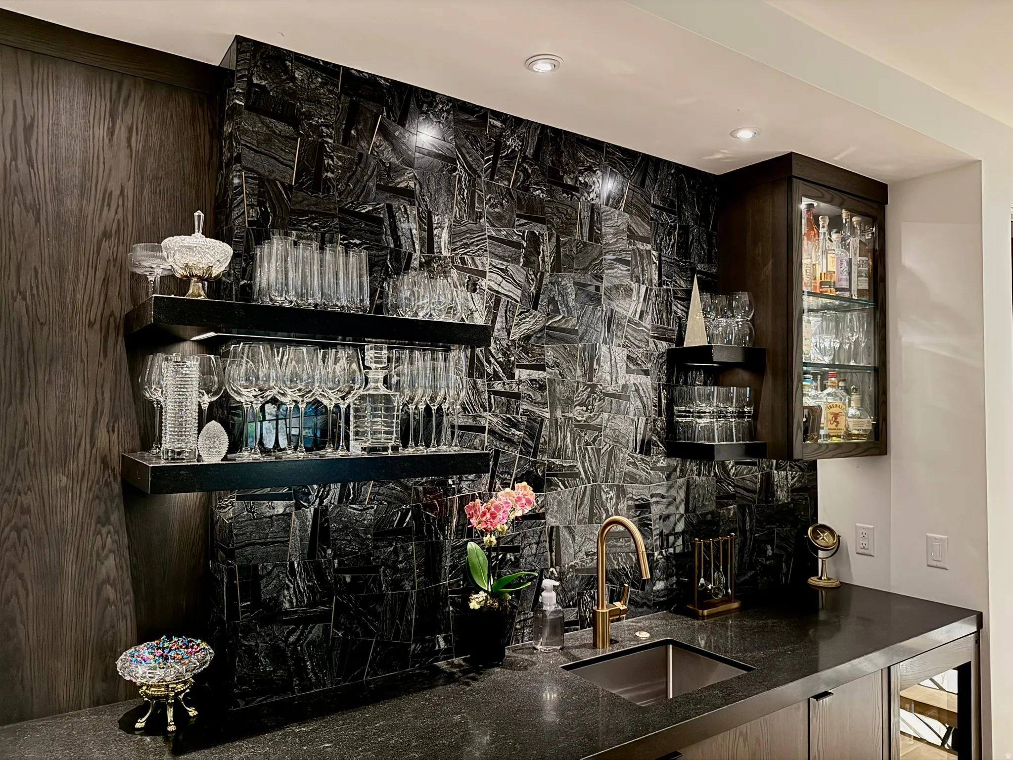 Built-in bar featuring a dark stone countertop, a square undermount sink with a brass-finish faucet, and a black tile backsplash